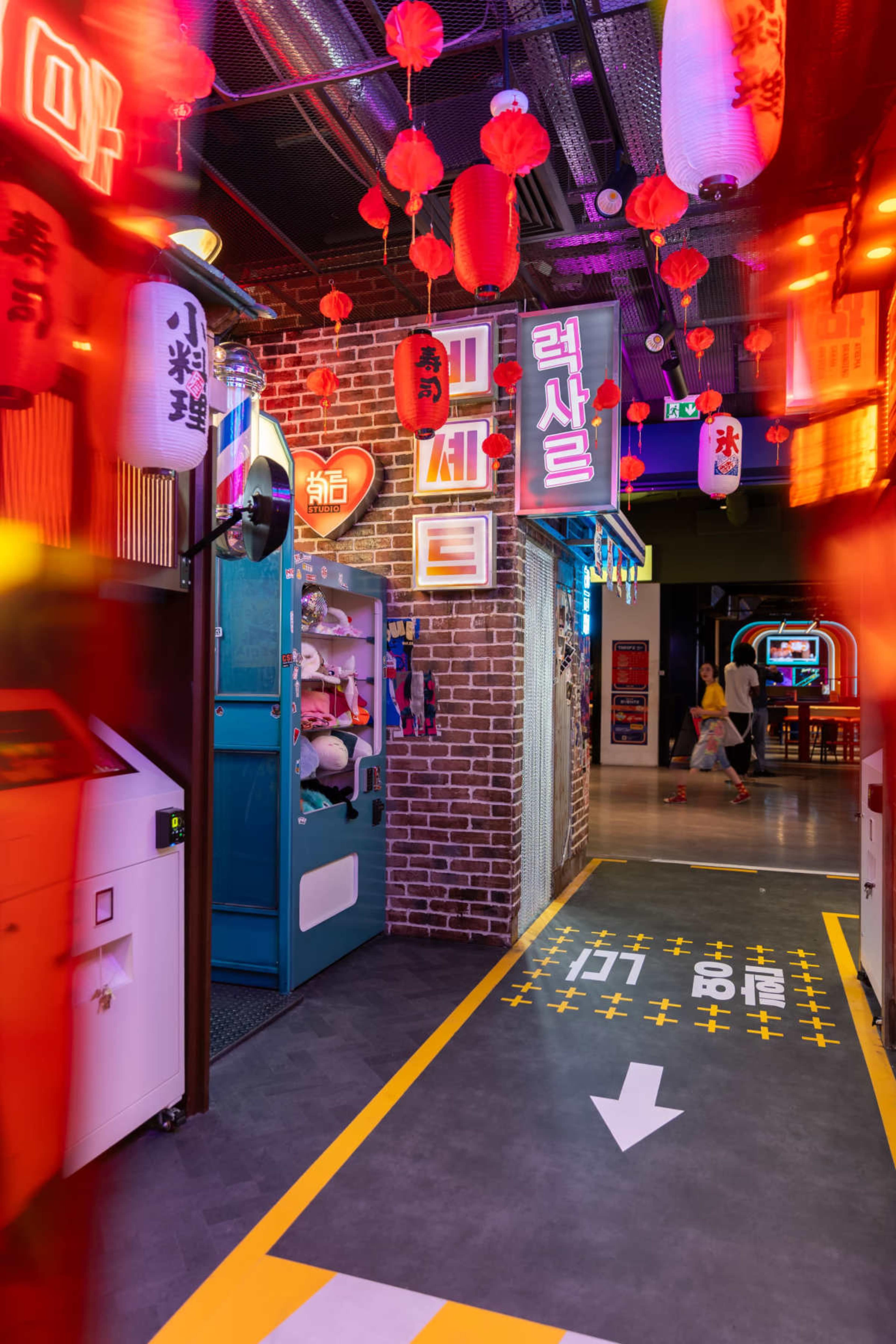The image depicts a vibrant hallway in an entertainment venue, lined with colorful neon signs and decorative lanterns, leading to various interactive booths and attractions.