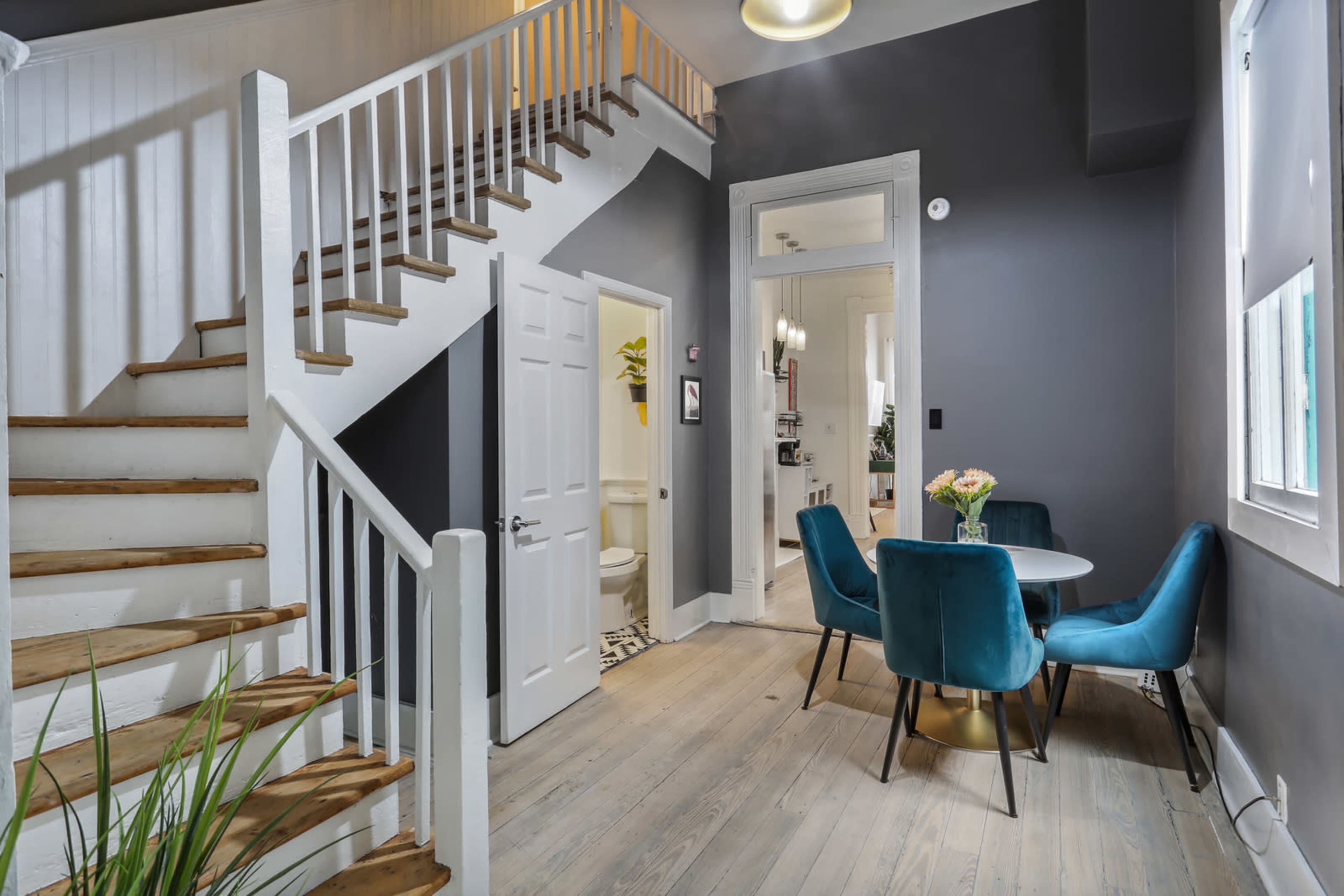 The image shows a modern interior featuring a staircase, a small dining area with a round table and blue chairs, and two doorways leading to other rooms.
