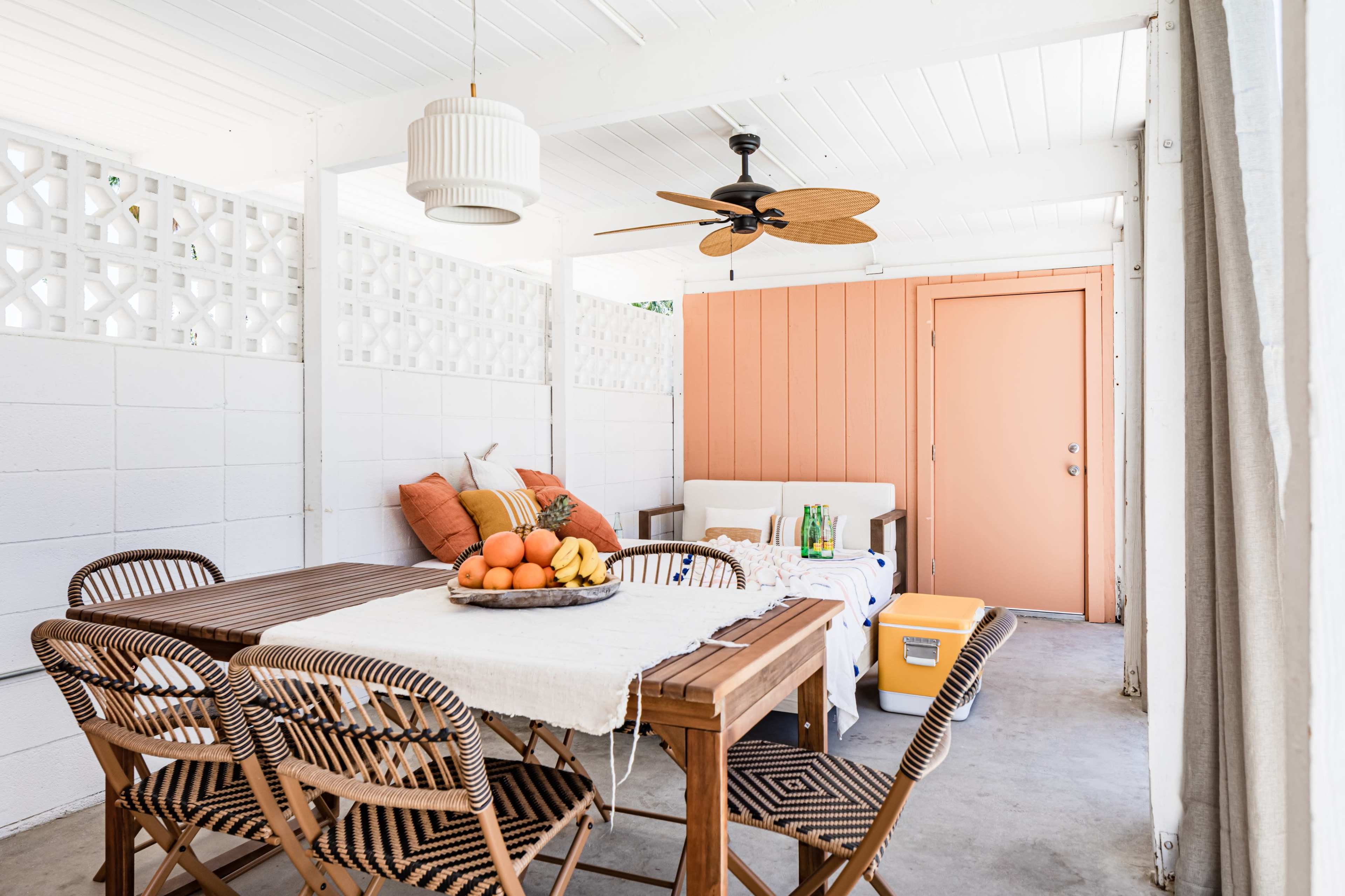 A covered outdoor area features a wooden dining table surrounded by woven chairs, a seating area with orange cushions, and a fan overhead.