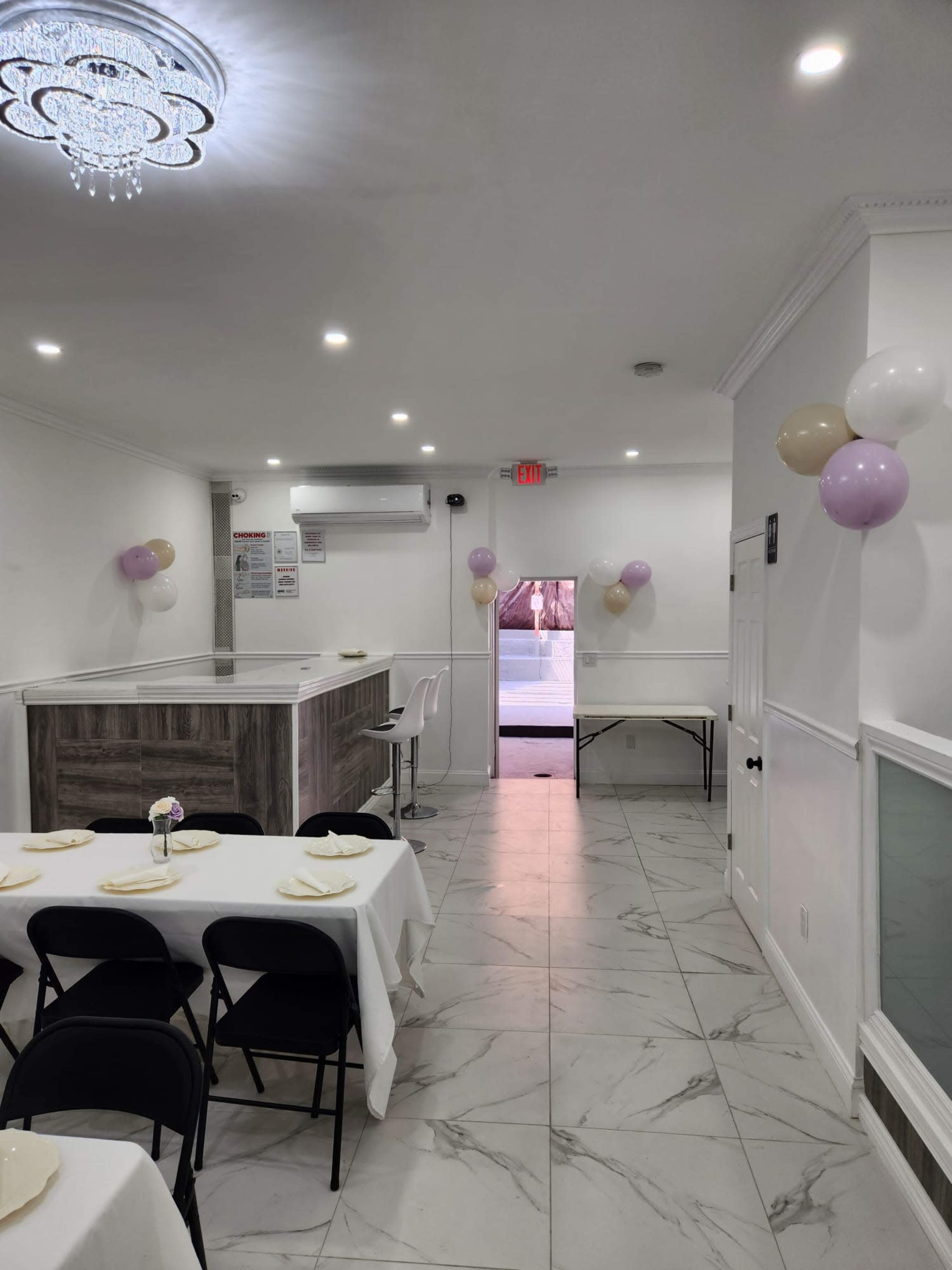 A brightly lit room with white walls, a wooden counter, tables set with white tablecloths, and balloons in the corners, leading to an open doorway.