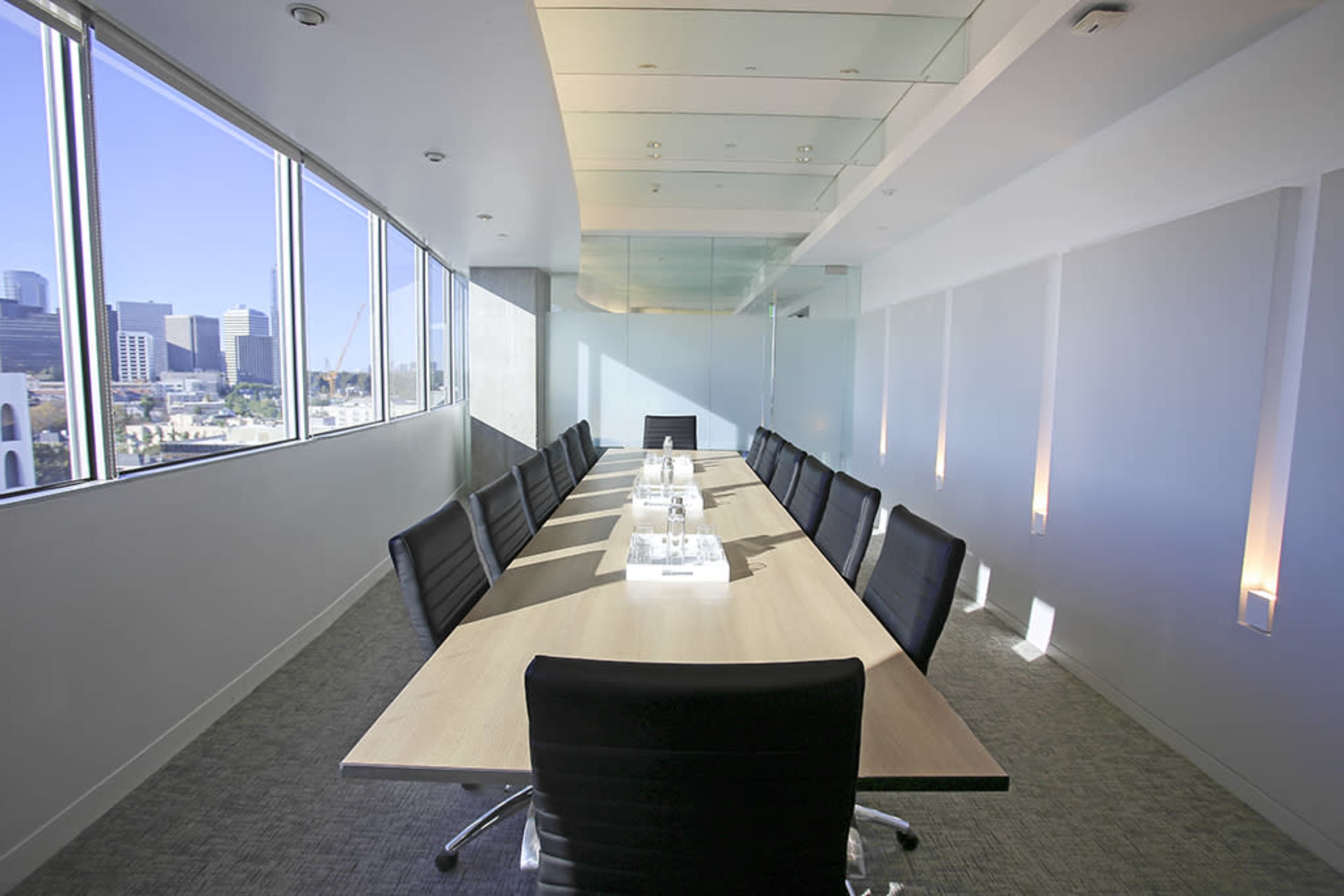 A modern conference room features a long wooden table surrounded by black chairs, with large windows offering a view of the city skyline.