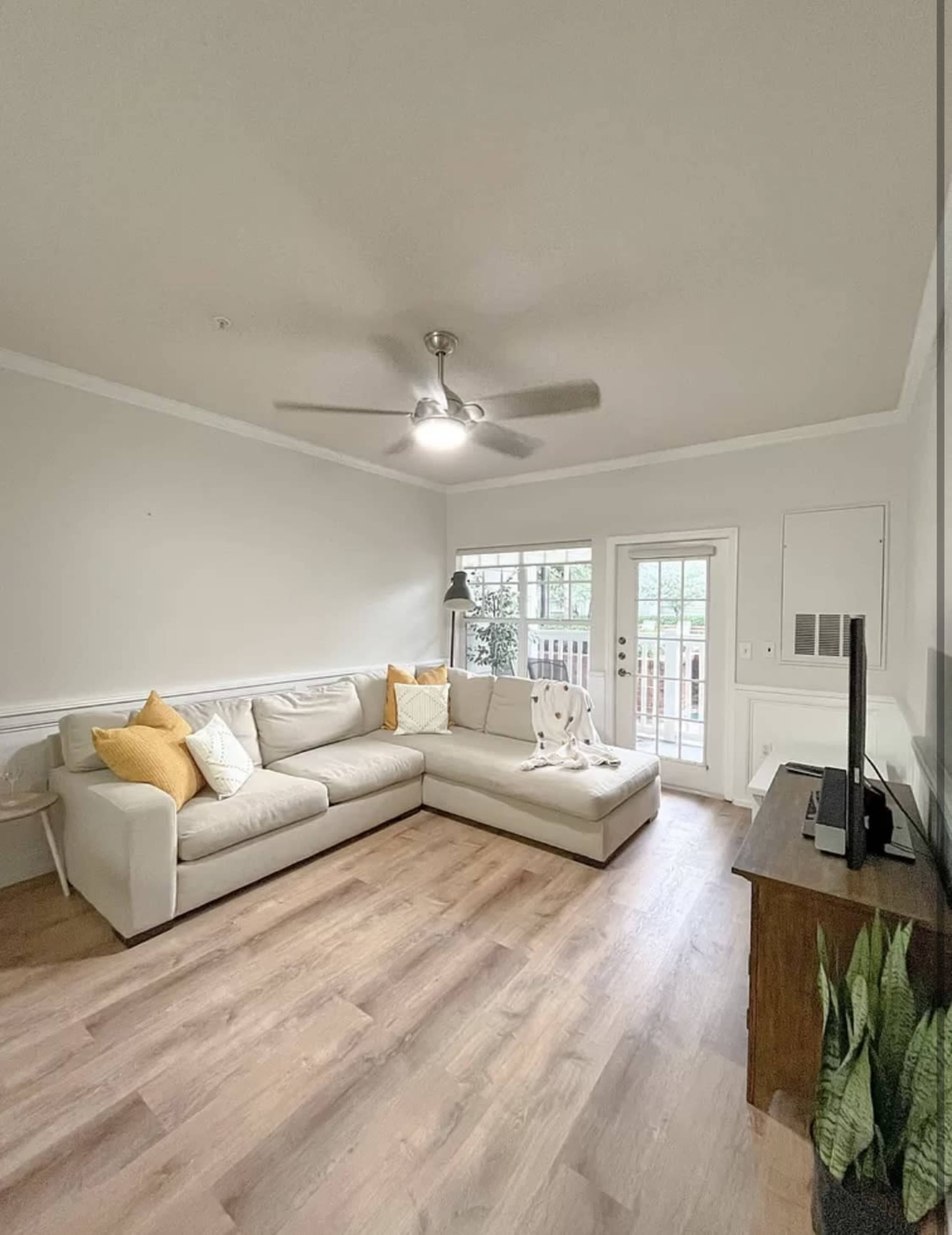 The image shows a living room featuring a light-colored sectional sofa, a wooden TV stand, and a view of sliding glass doors leading to a patio.