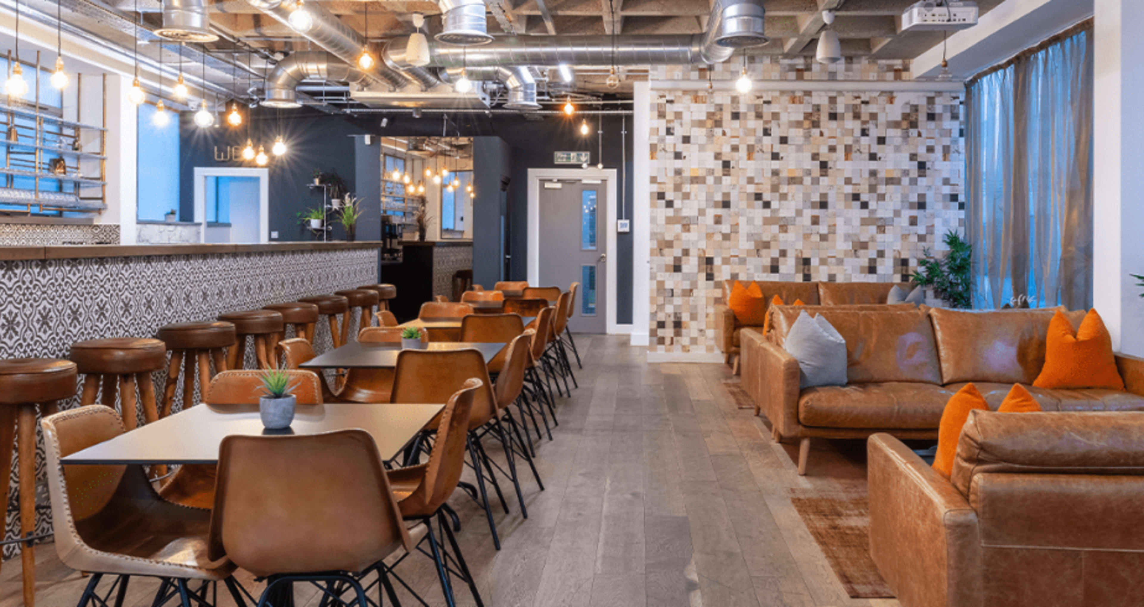 A modern lounge area featuring leather seating, wooden tables, and a patterned wall, with warm lighting and a bar in the background.