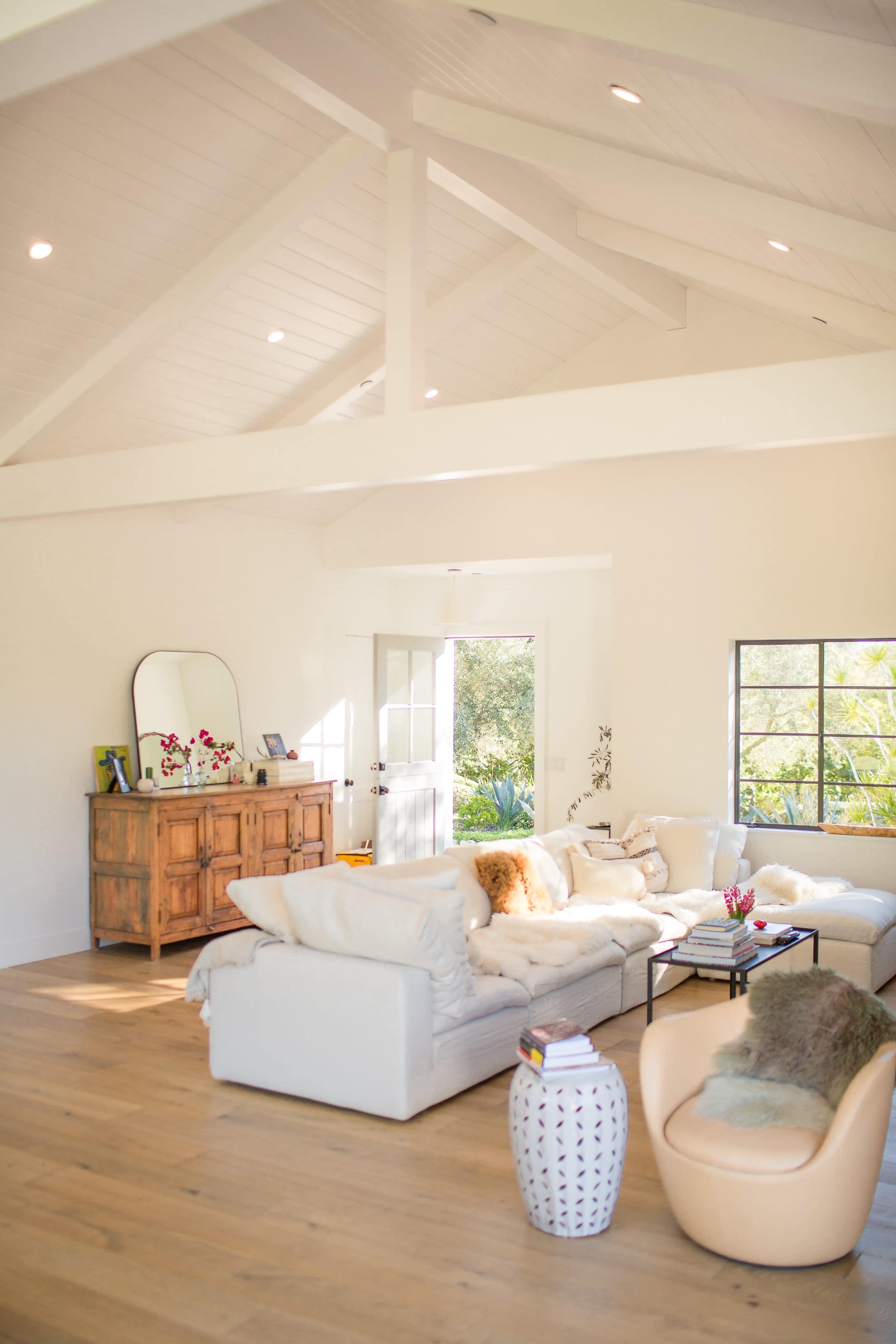 A bright, airy living room features a white couch, wooden sideboard, and large windows that allow natural light to fill the space.