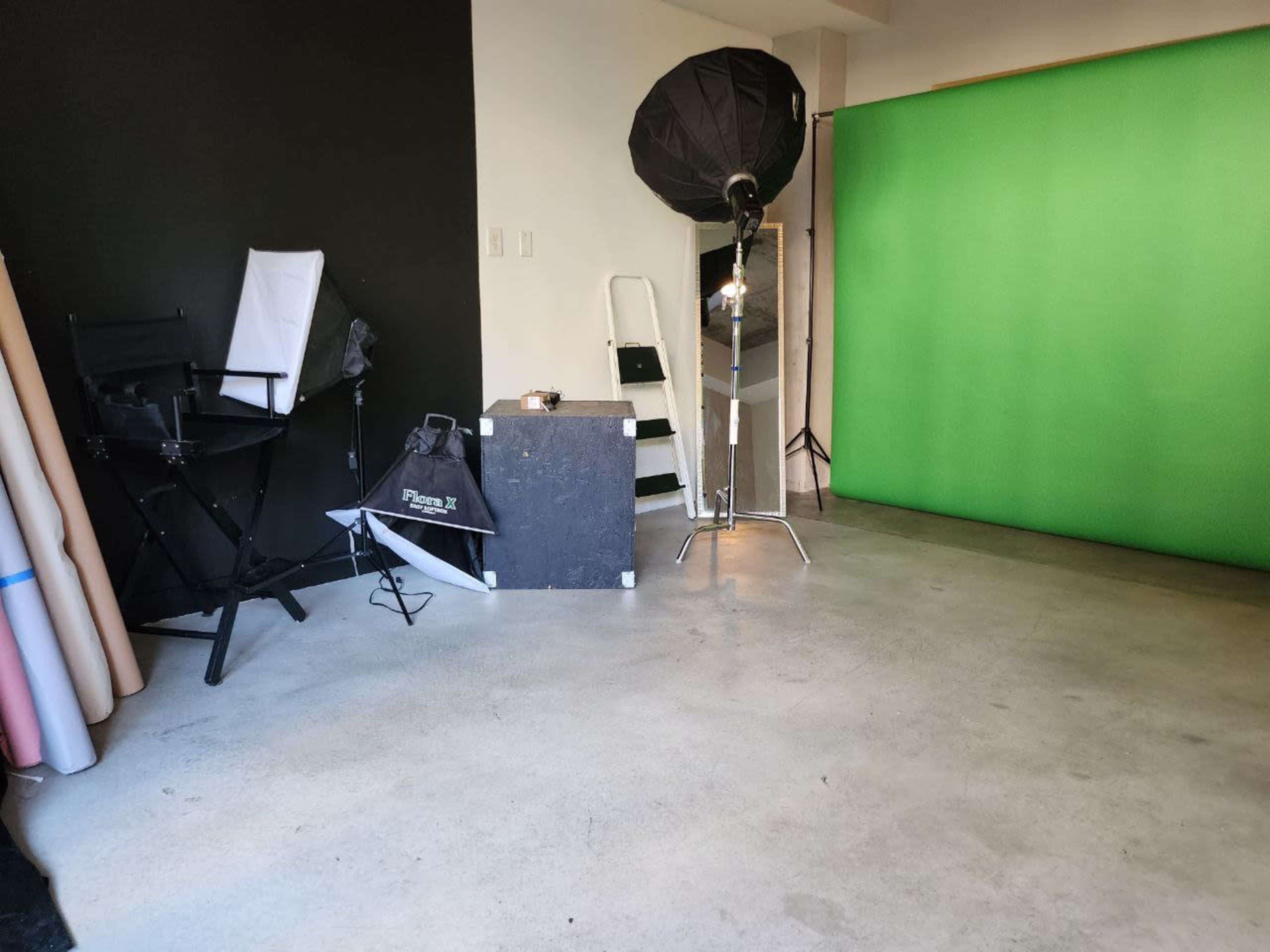 The image shows a photography studio setup with a green backdrop, lighting equipment, a folding chair, and various other studio supplies.