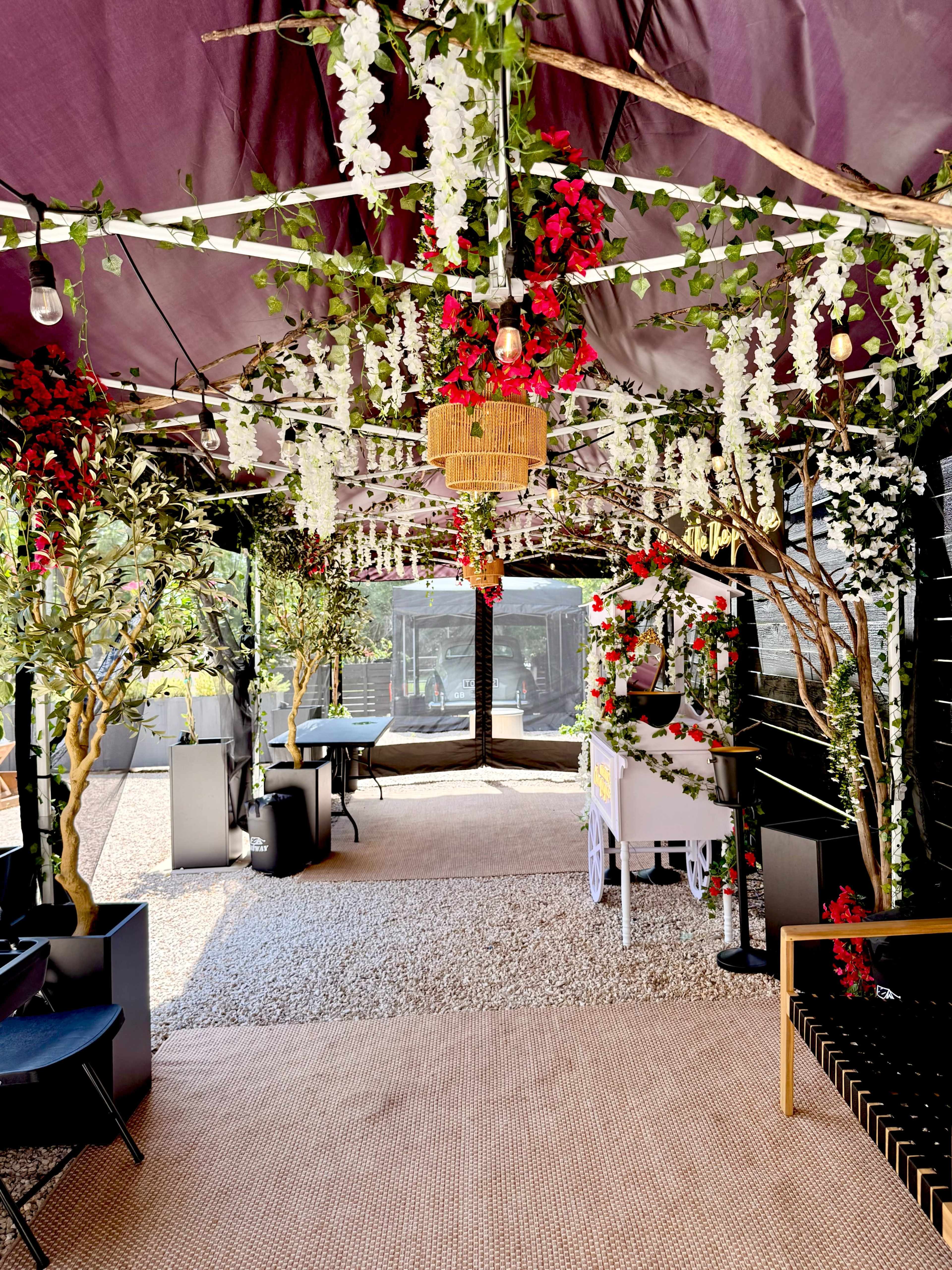 The image shows a tented space decorated with artificial flowers, greenery, and hanging lights, creating a vibrant outdoor dining atmosphere.