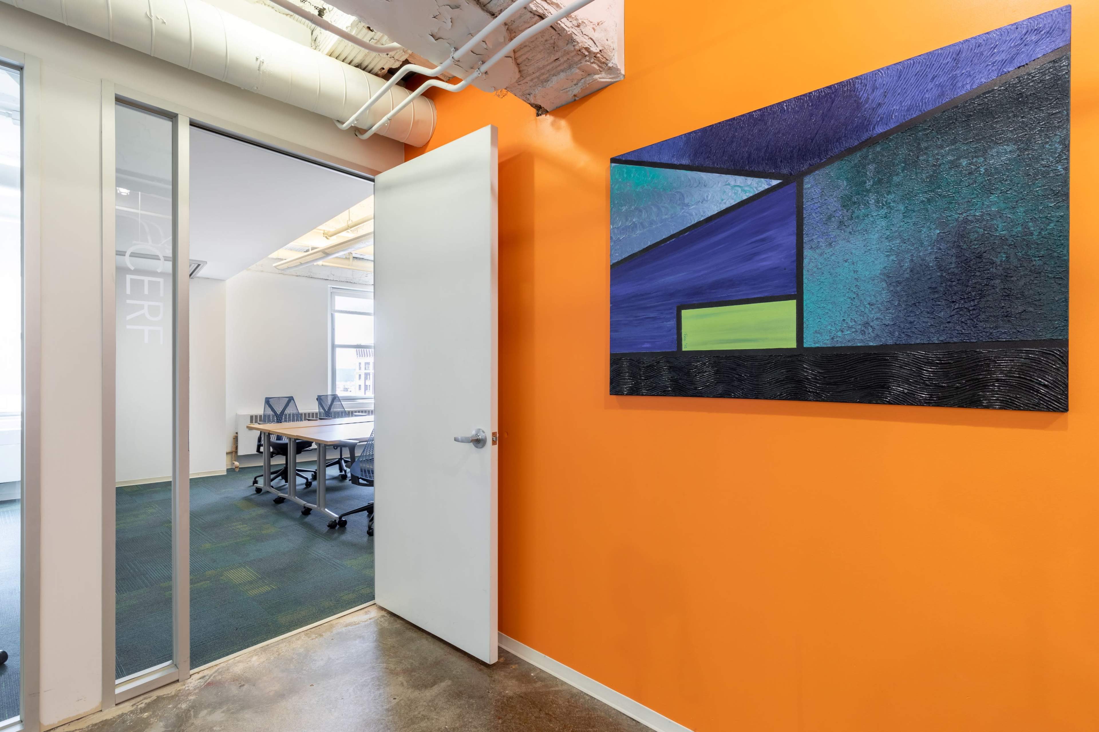 An orange wall features an abstract painting, while a doorway leads to a conference room with a table and chairs.