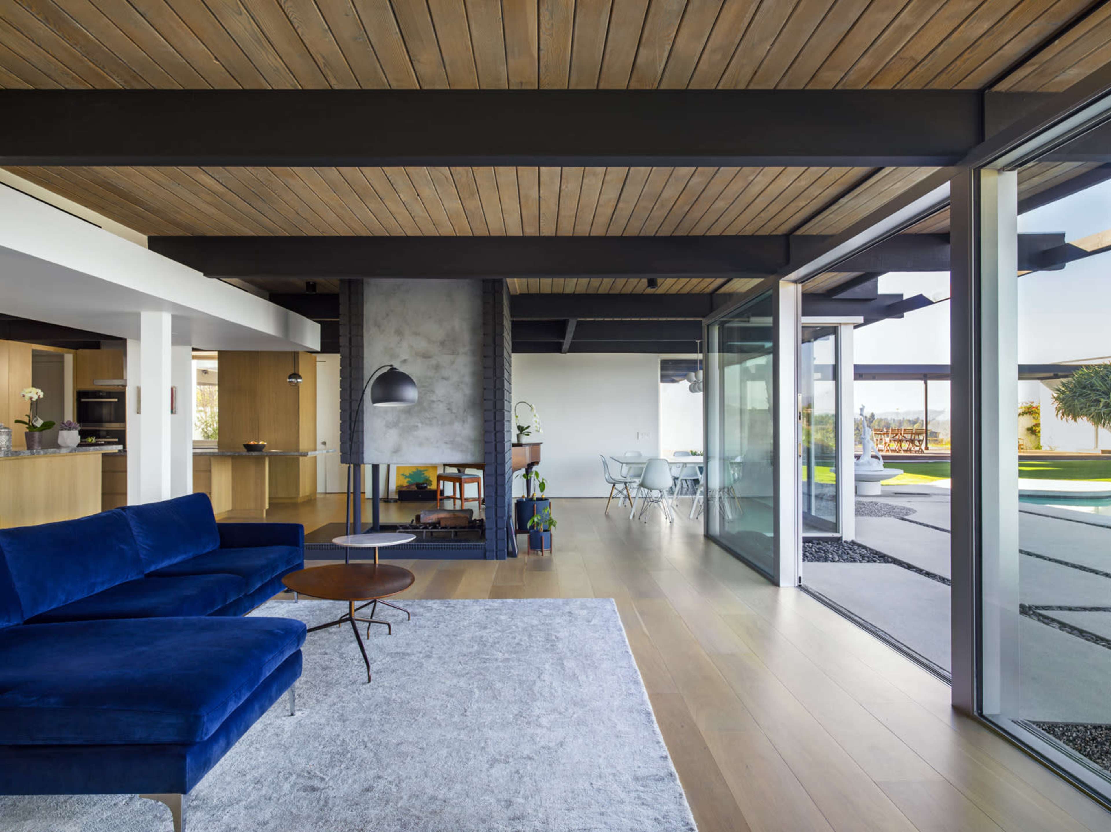 The image shows a modern living space featuring a blue sofa, a glass wall opening to a patio, and wooden ceiling beams.