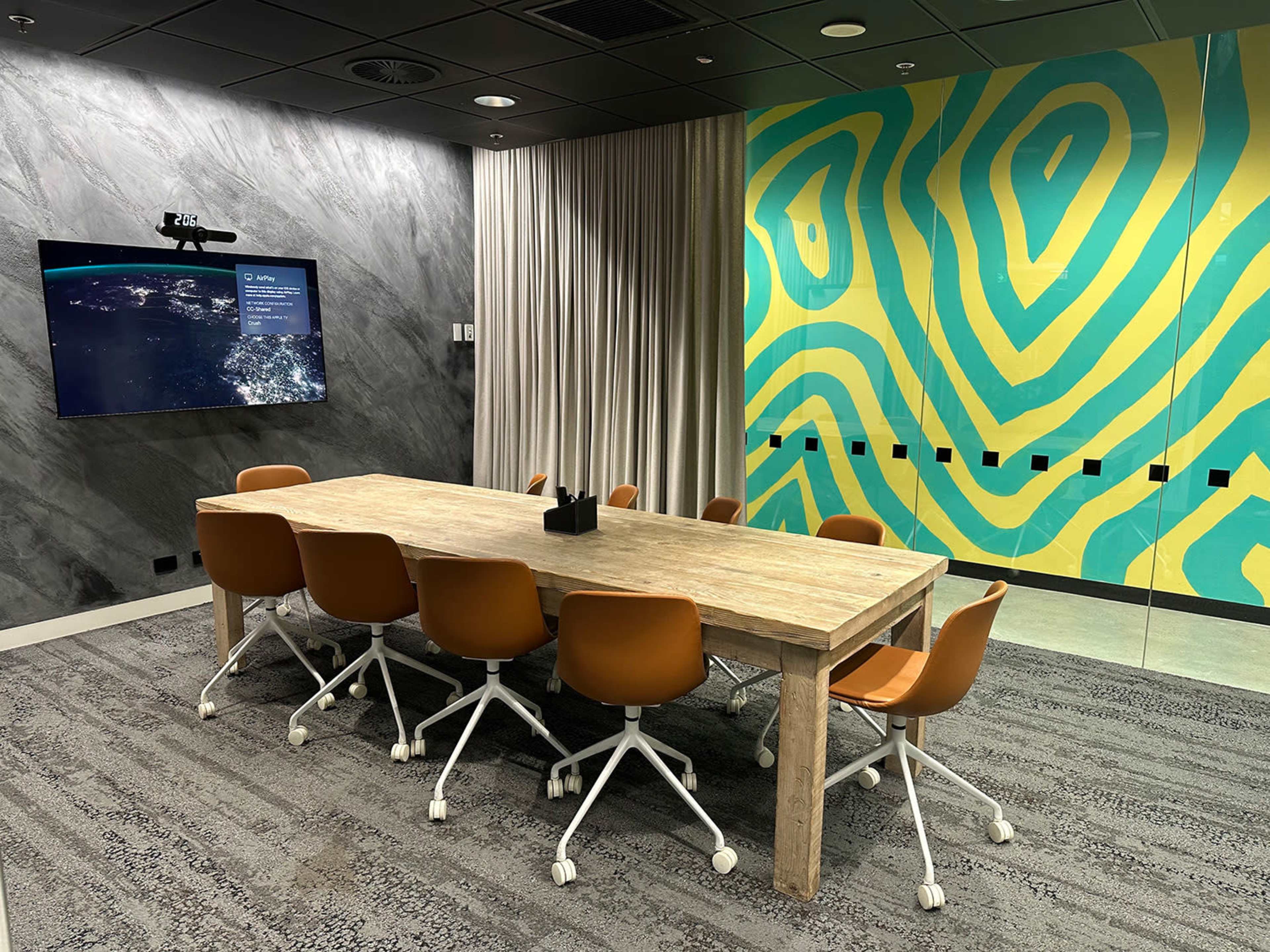 A conference room features a large wooden table surrounded by orange chairs, with a screen displaying information and a vibrant green and yellow patterned wall.