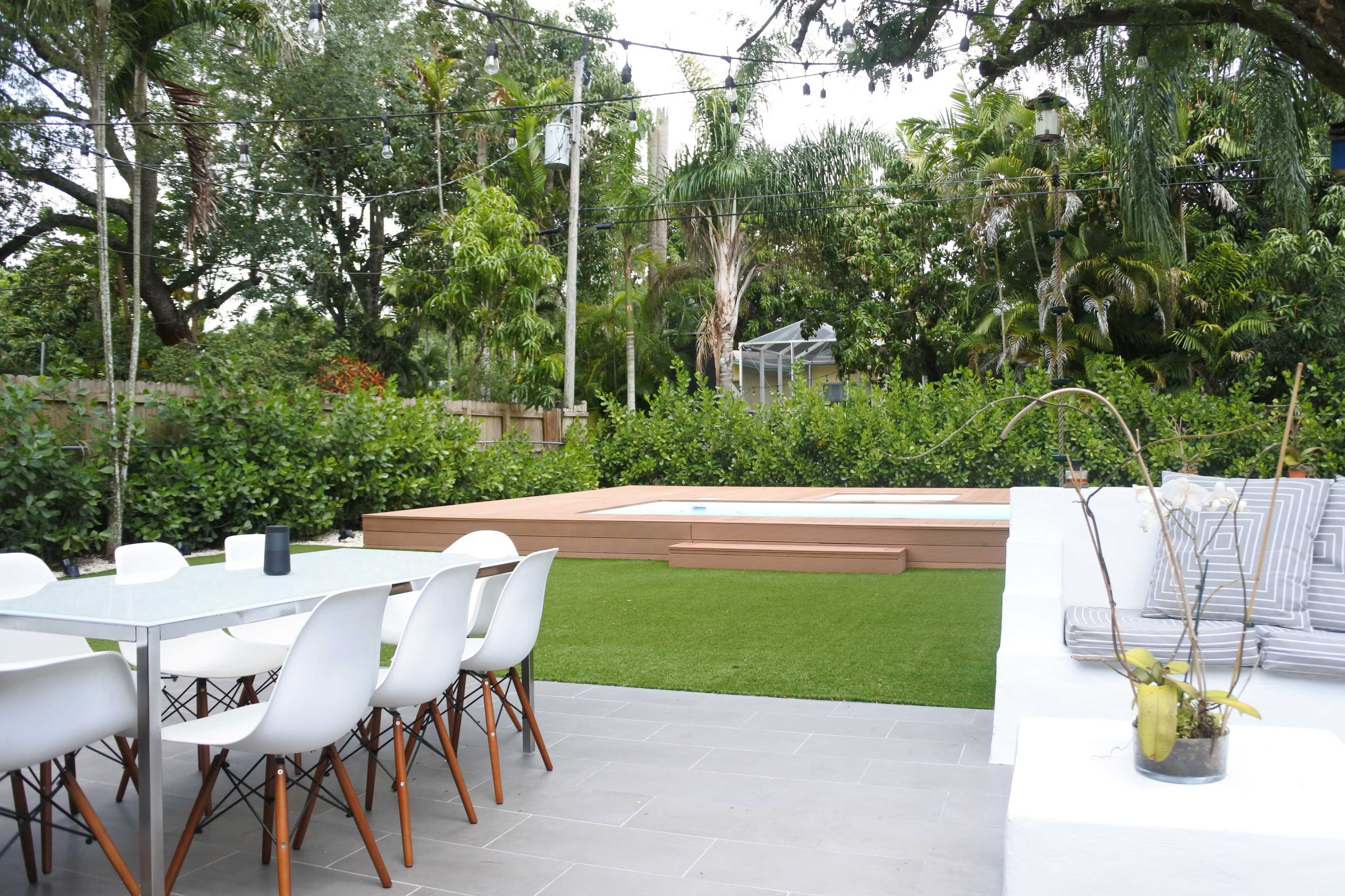 A modern outdoor patio features a dining table with white chairs, a small swimming pool, and lush greenery surrounding the area.