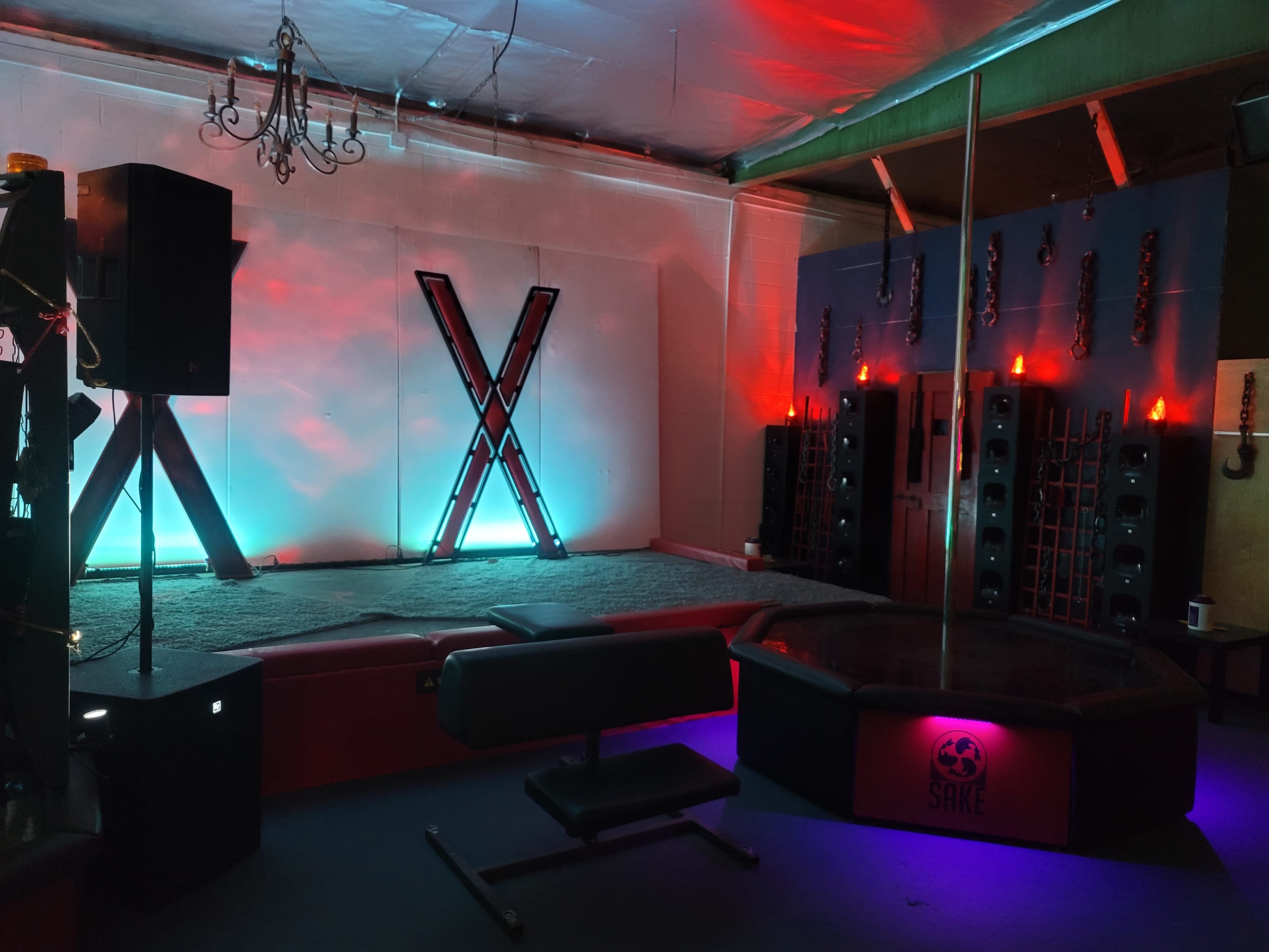 A dimly lit room designed for performances, featuring a pole in the center, large speakers along the walls, and decorative lighting.