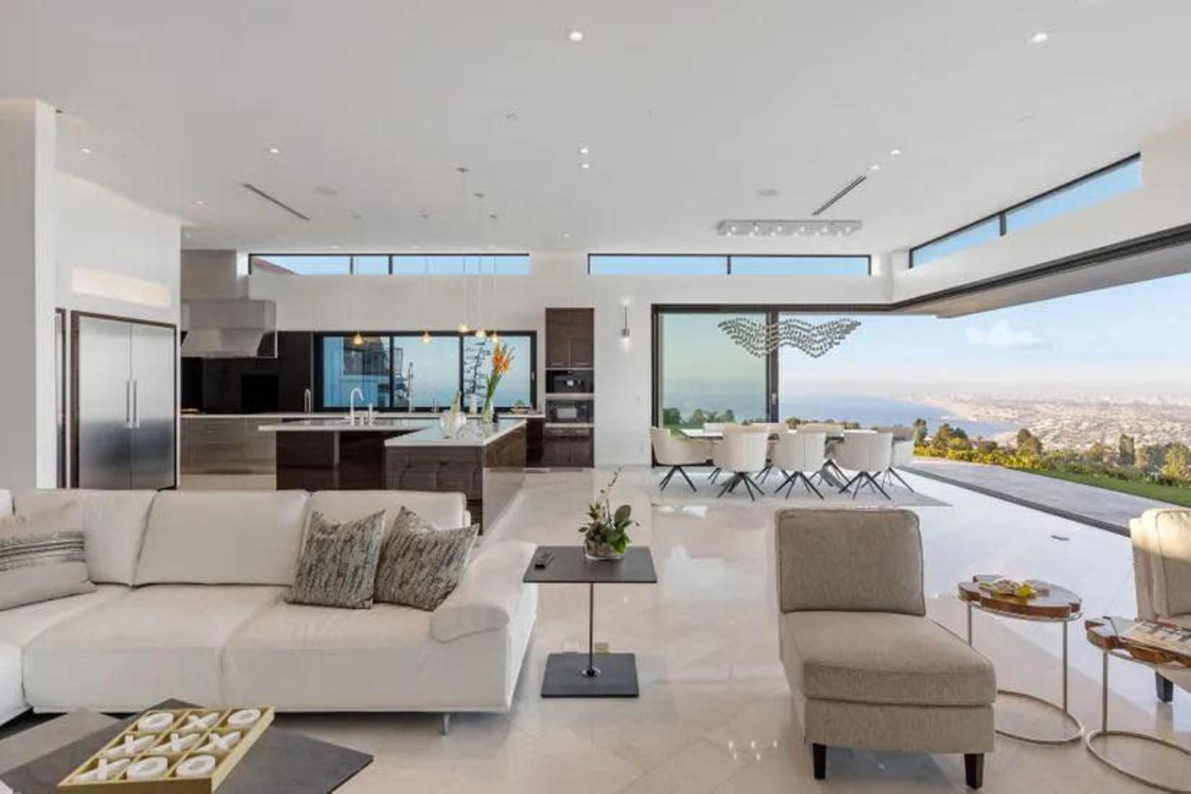 A modern living room with large windows offering a panoramic view, featuring a white sofa, a minimalist coffee table, and a dining area with a sleek design.