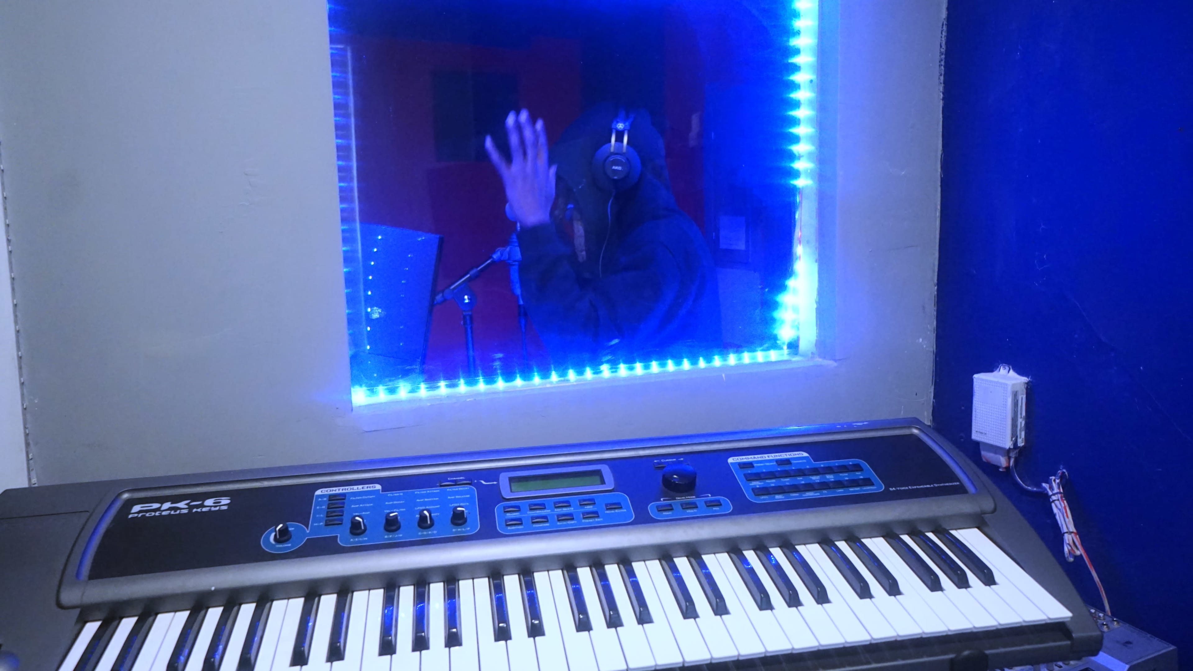A musician wearing headphones stands behind a glass partition in a recording studio, while a keyboard rests in the foreground.