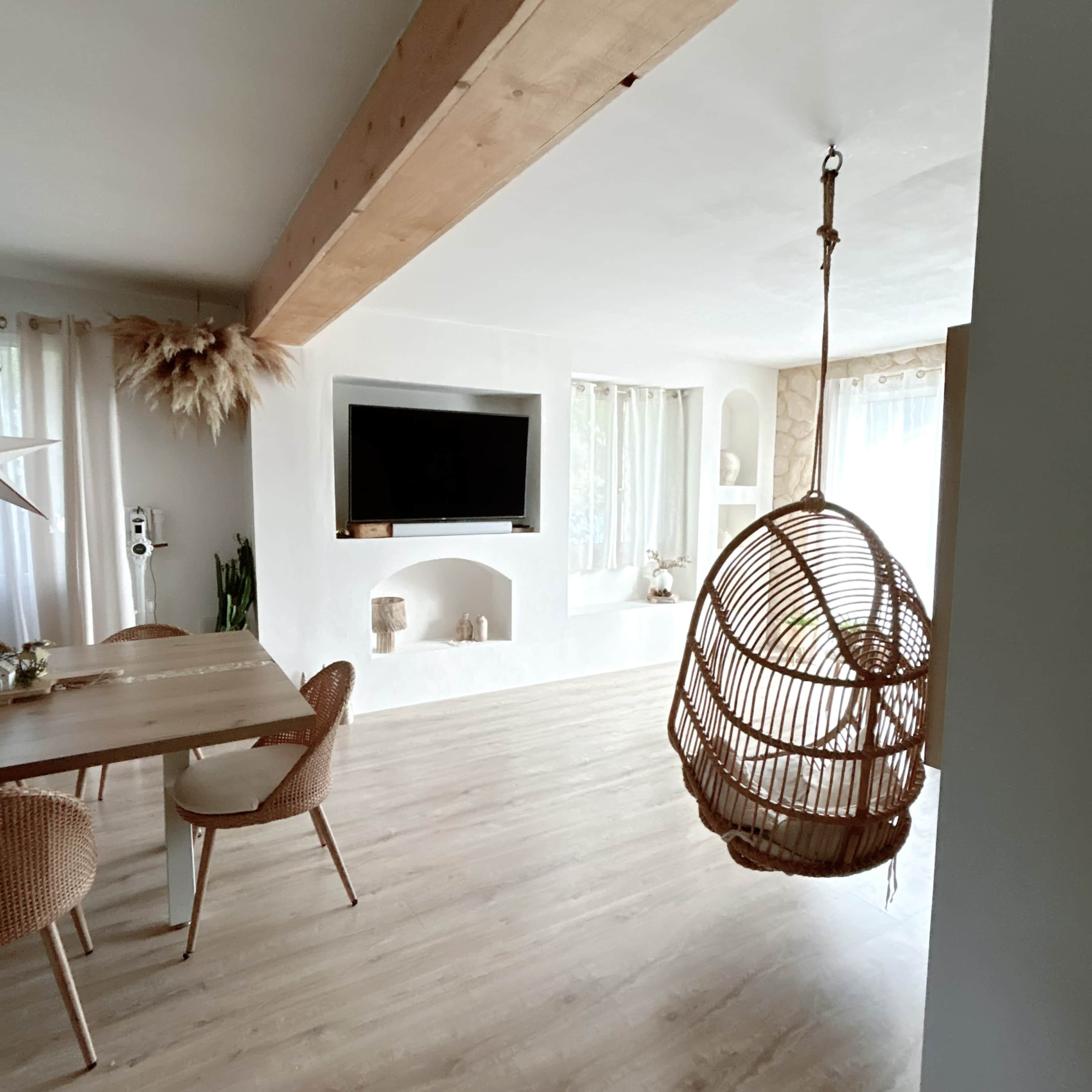 A spacious living area features a wooden beam ceiling, a hanging rattan chair, and a dining table surrounded by woven chairs, all under soft natural lighting.