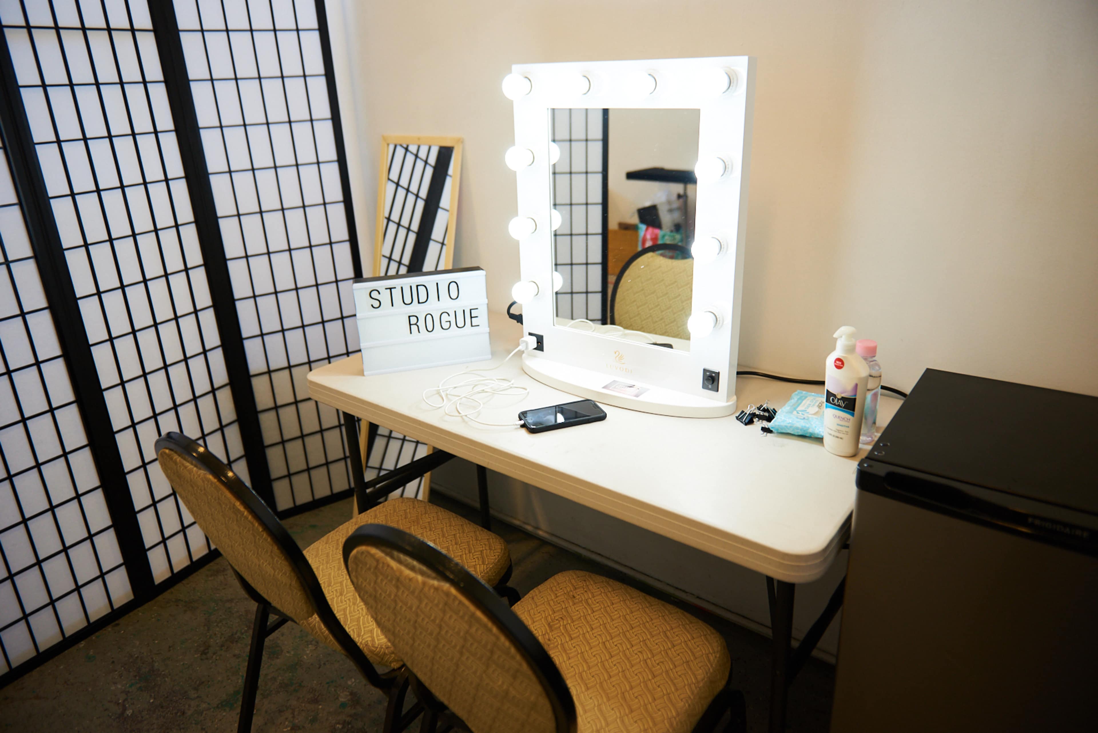 A well-lit vanity mirror with bulbs is positioned on a table alongside a sign that reads "STUDIO ROGUE," two chairs, and a small refrigerator in a room with partition screens.