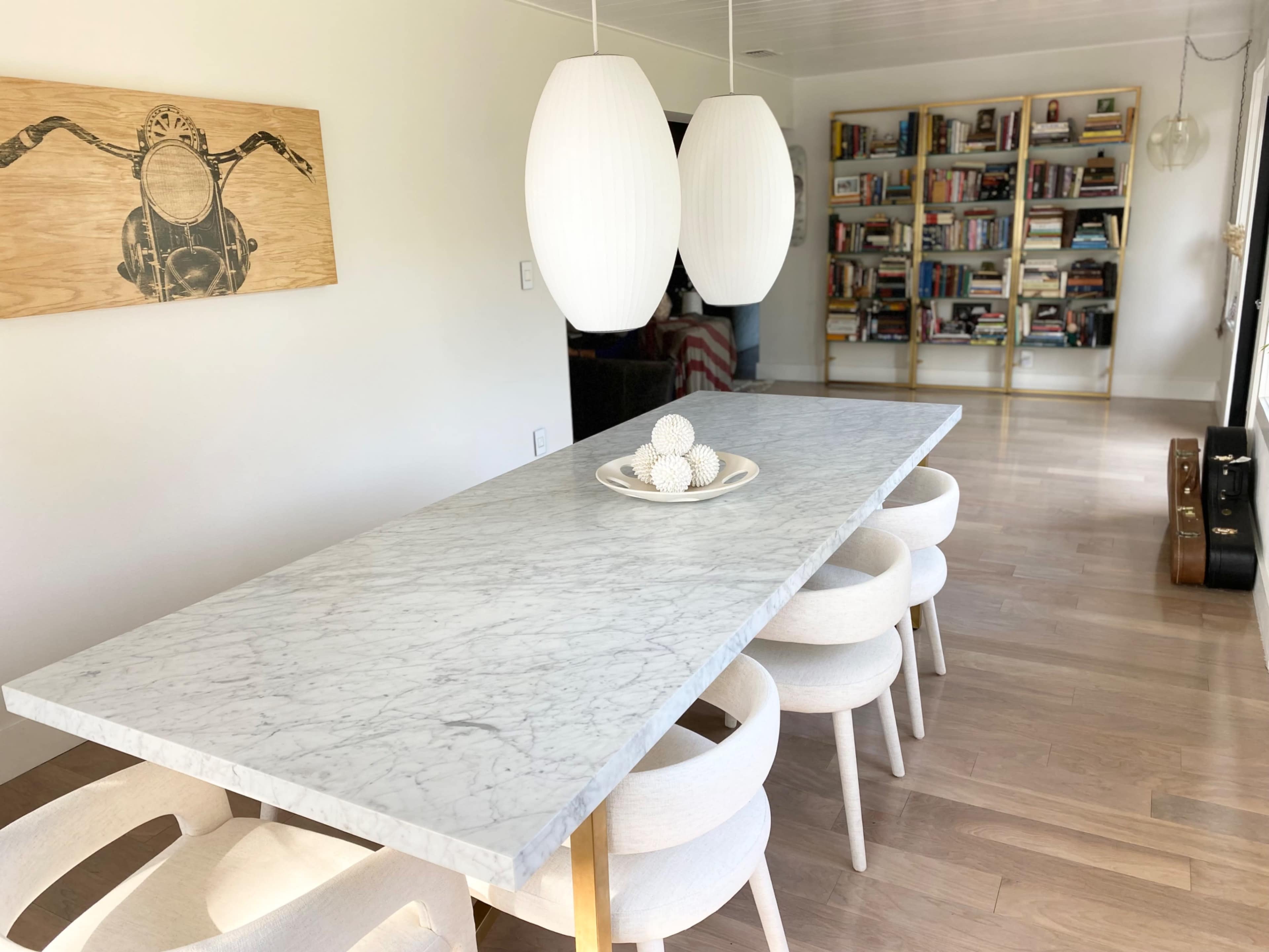 A long marble dining table with white chairs is positioned in a bright room, next to a bookshelf filled with various books.