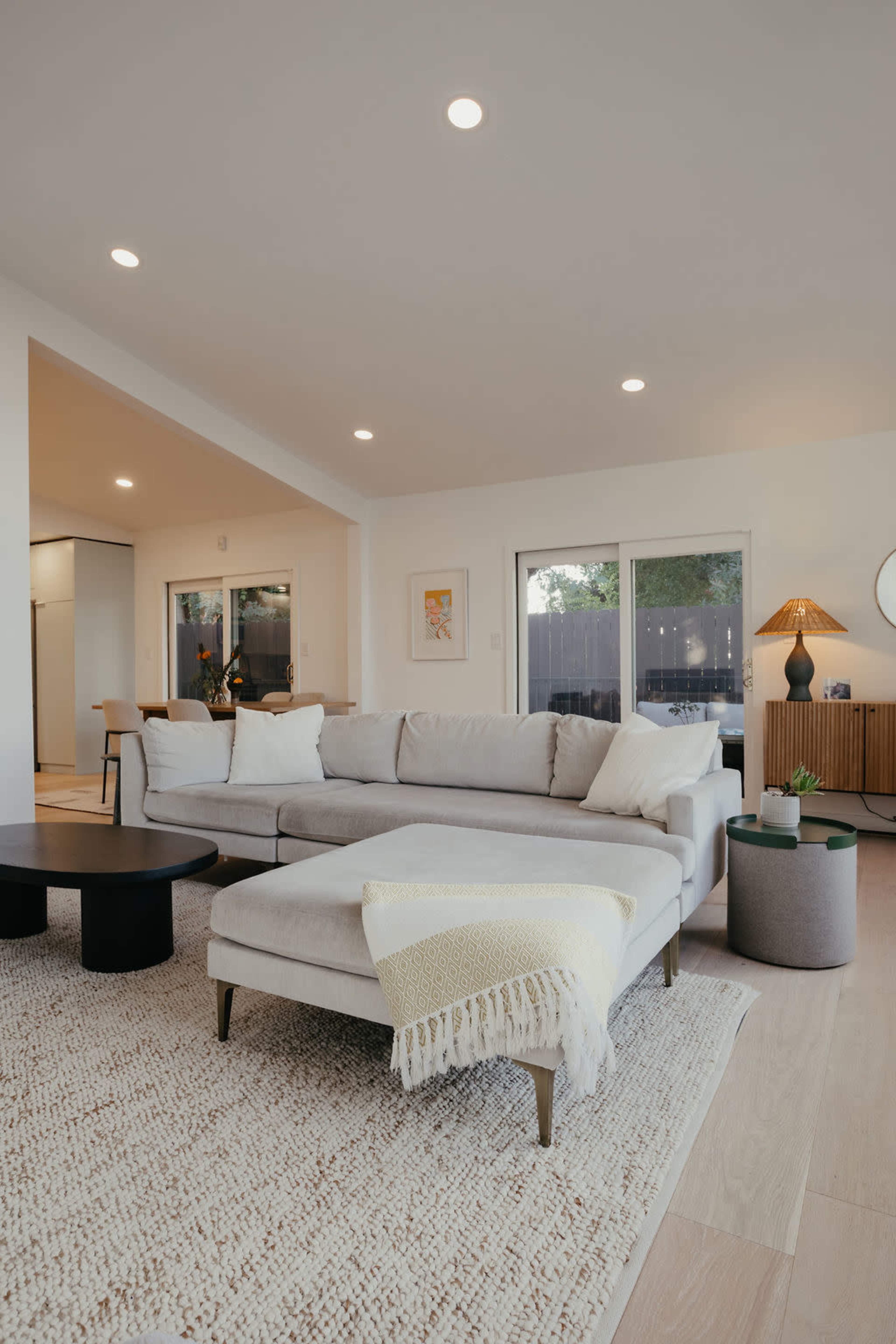 A modern living room features a light gray sectional sofa, a round black coffee table, and large windows allowing natural light to fill the space.