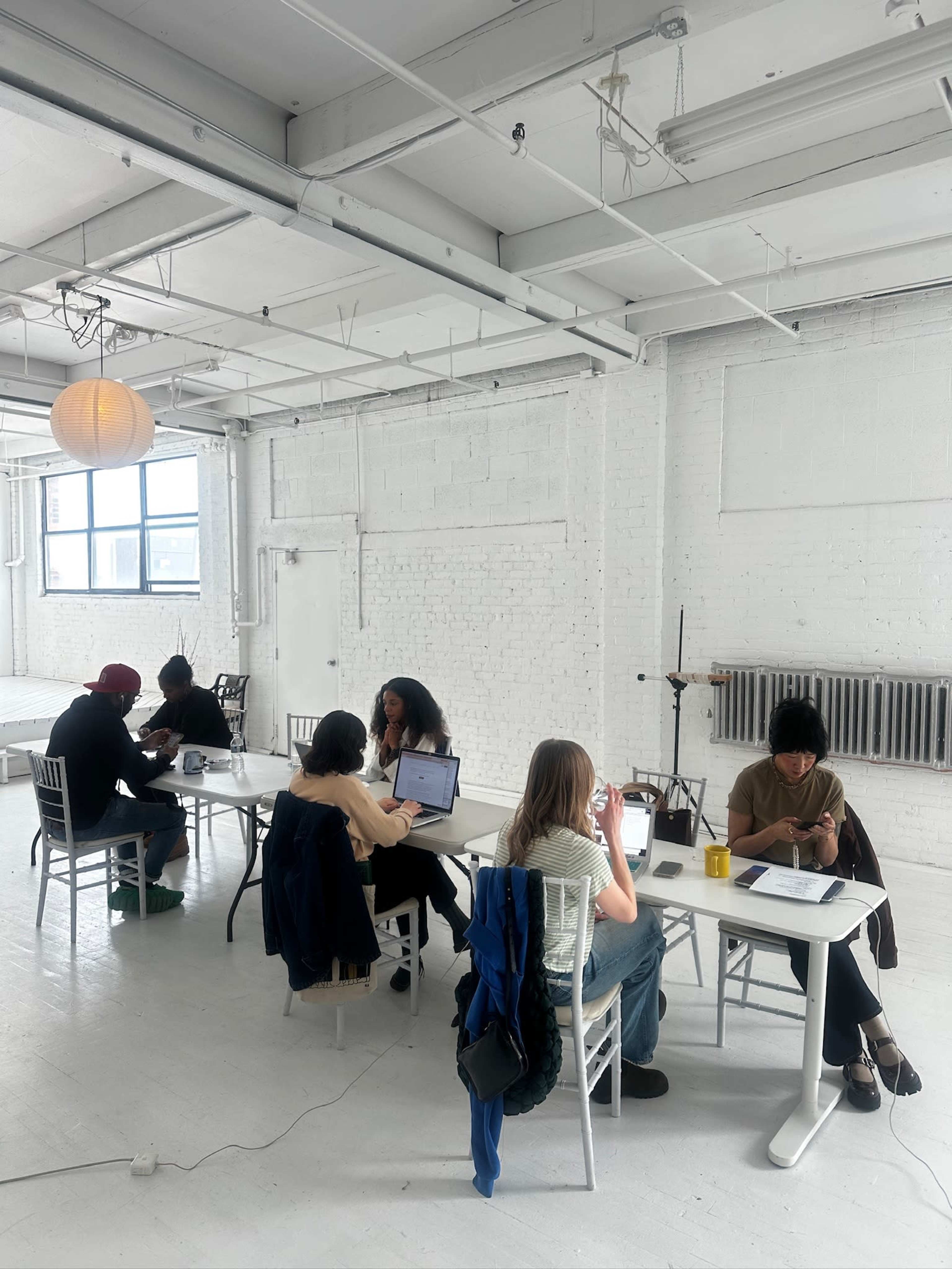A group of people are seated at tables in a bright, airy room with white walls and large windows, engaged in various tasks on laptops and mobile devices.