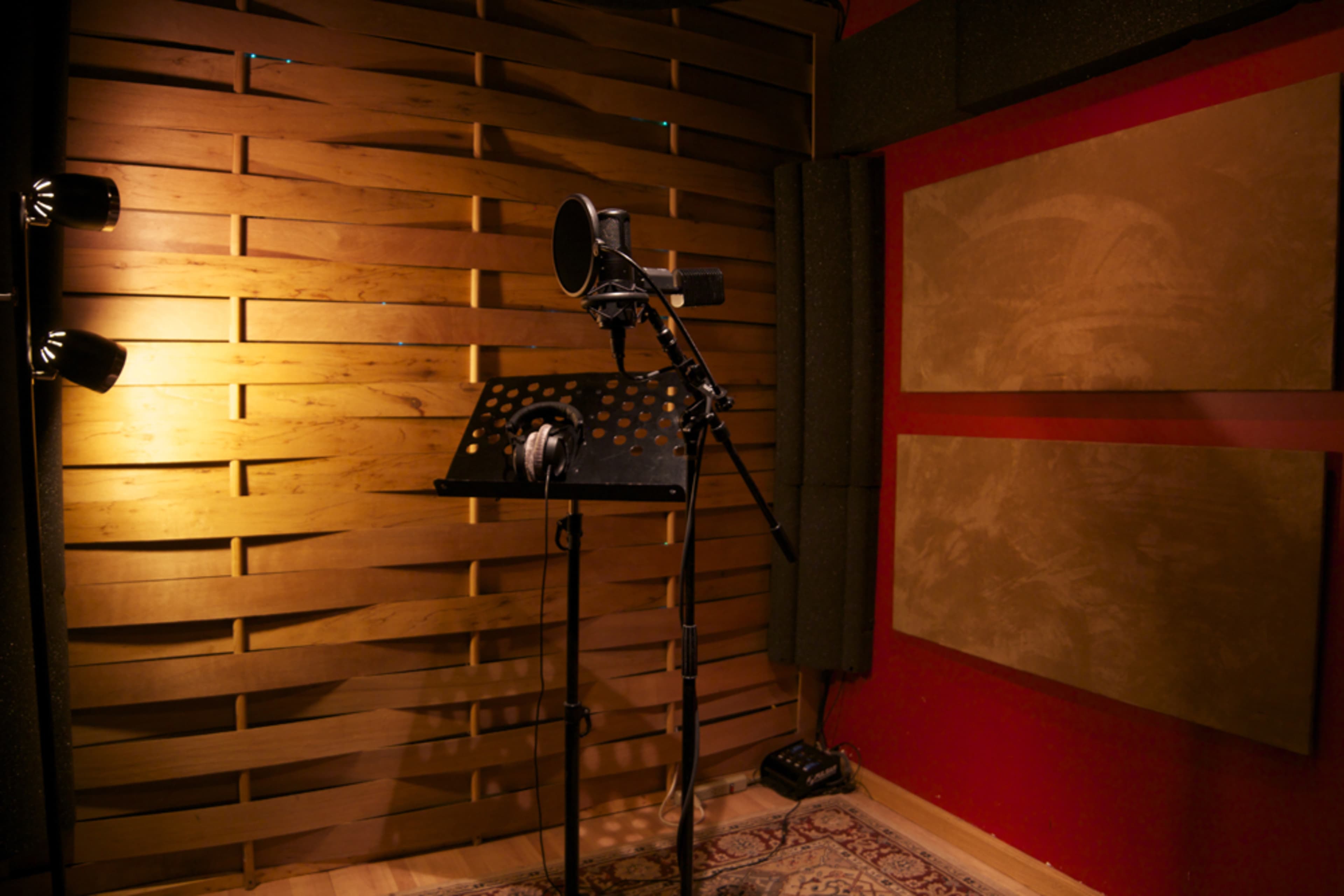 The image shows a recording studio with a microphone on a stand, headphones hanging on a music stand, and wooden paneling on the walls.