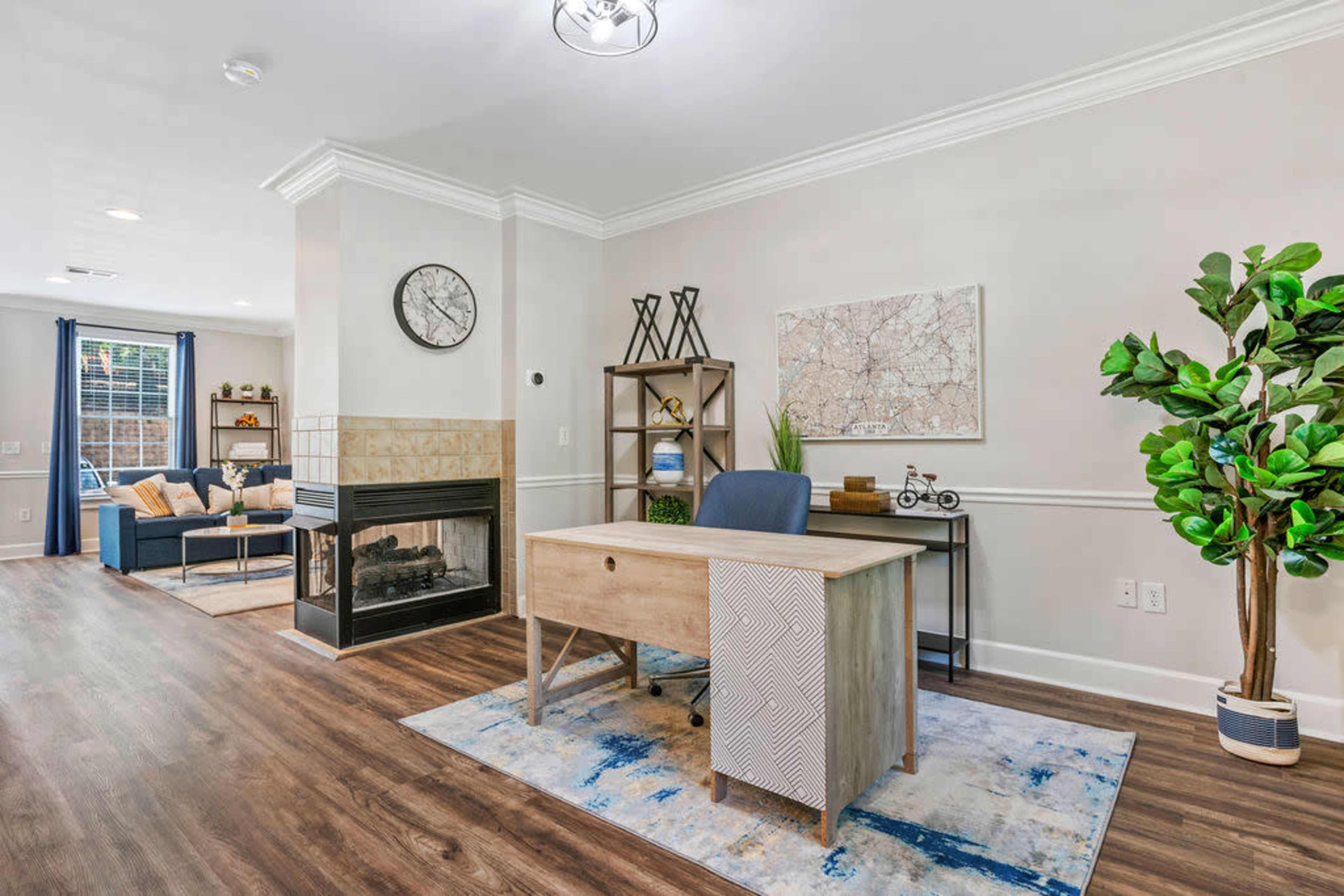 A modern home office features a wooden desk and office chair in front of a fireplace, with a living area visible in the background.