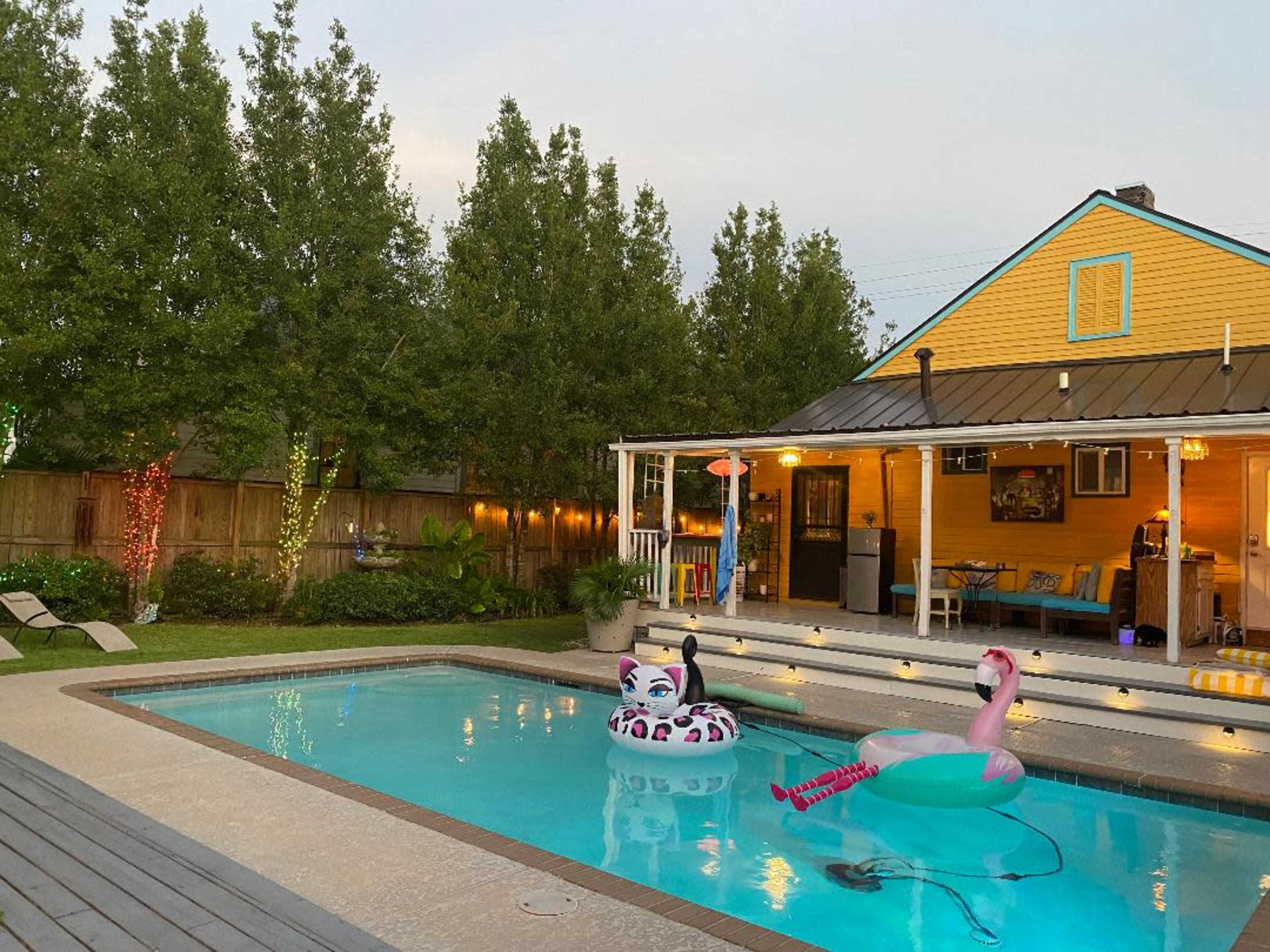 A backyard features a swimming pool with colorful inflatable floats and a yellow house adorned with string lights and a patio.