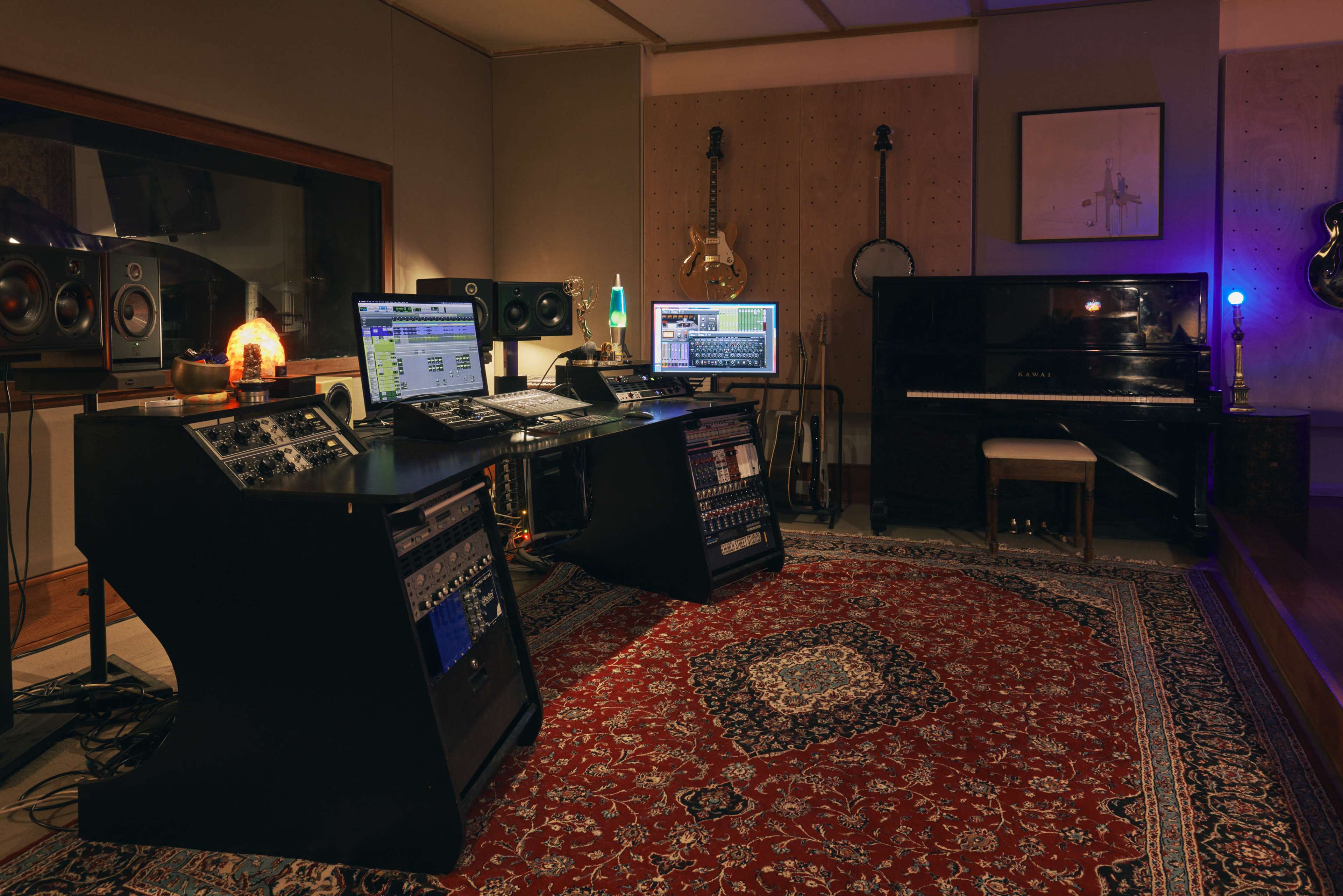 The image shows a music studio equipped with audio equipment, computers, and a grand piano, all arranged on a patterned rug.
