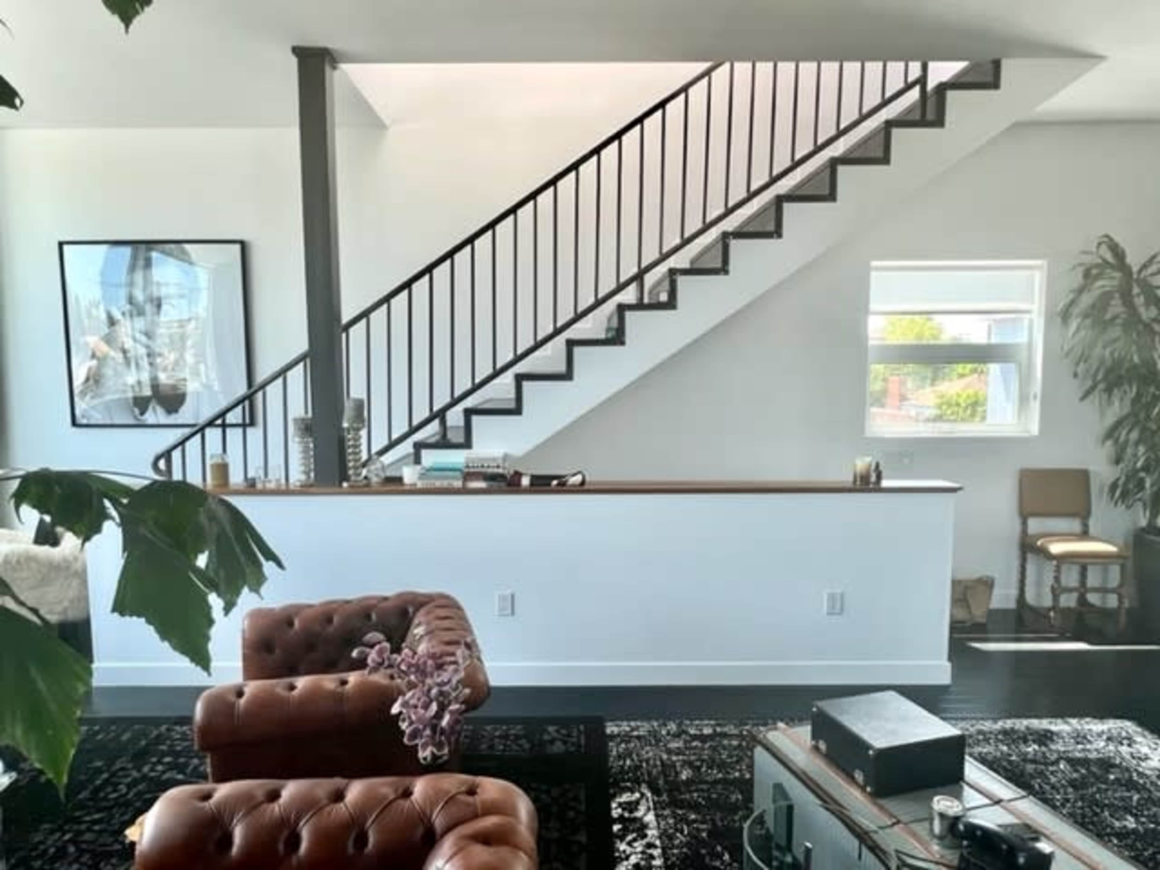 A spacious living room features a leather couch, a staircase against the wall, and large windows letting in natural light.
