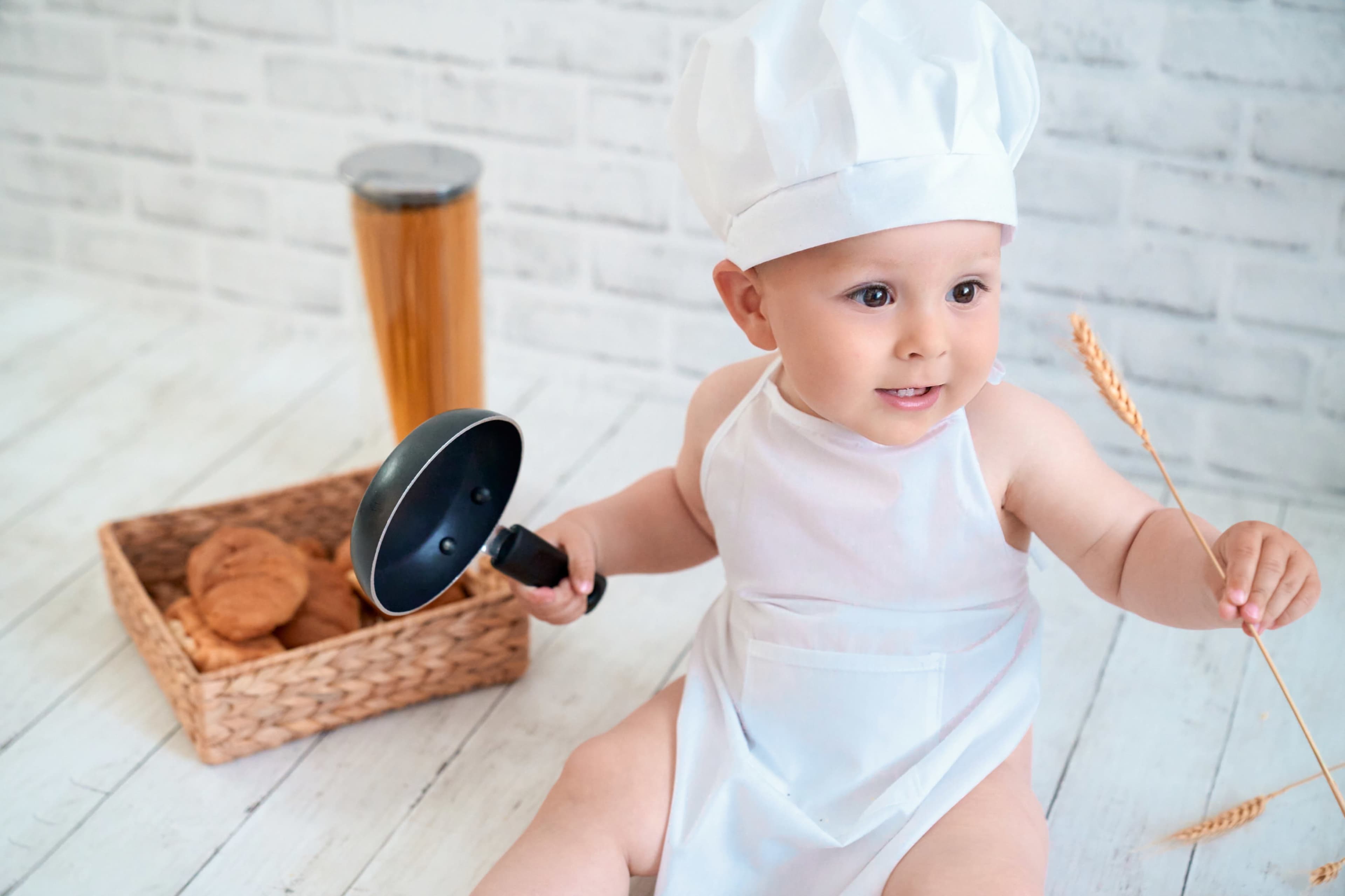 A baby wearing a chef's hat and an apron is sitting on the floor, holding a small frying pan in one hand and a piece of wheat in the other, with a basket of cookies nearby.