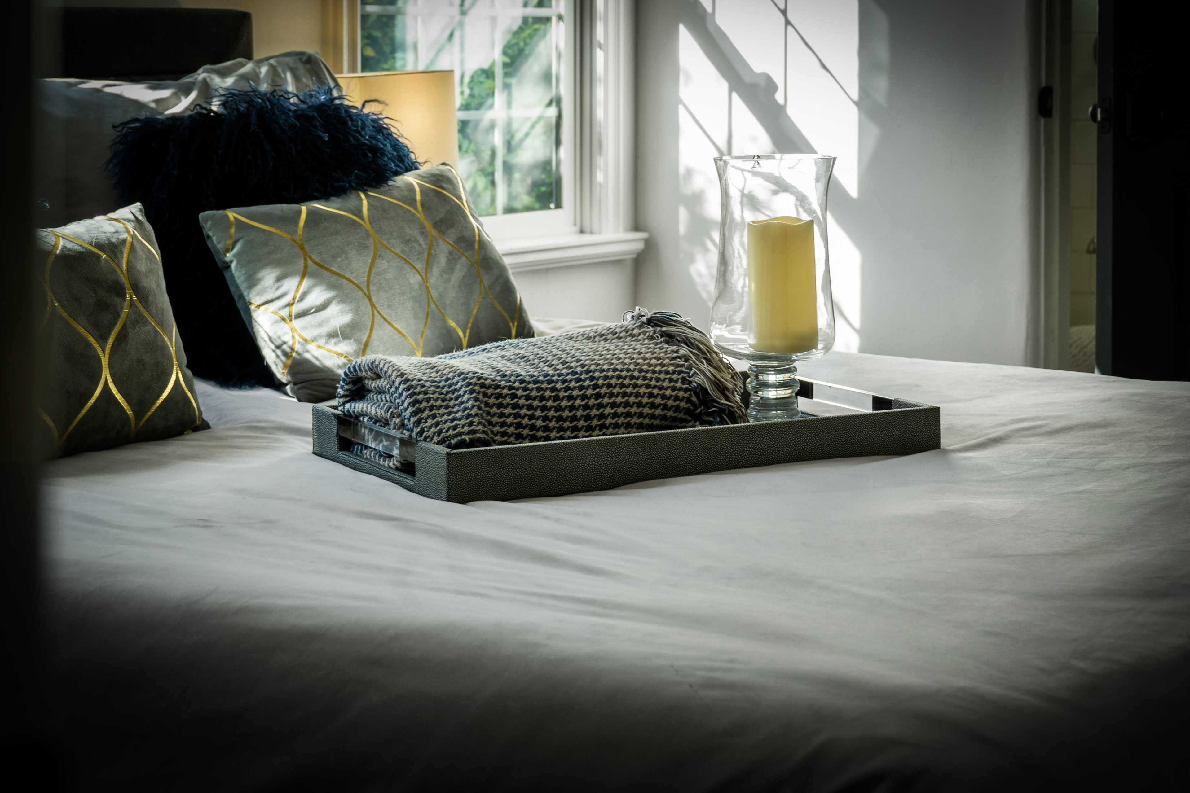 A neatly arranged tray with a candle and a folded blanket rests on a bed adorned with decorative pillows in a well-lit room.