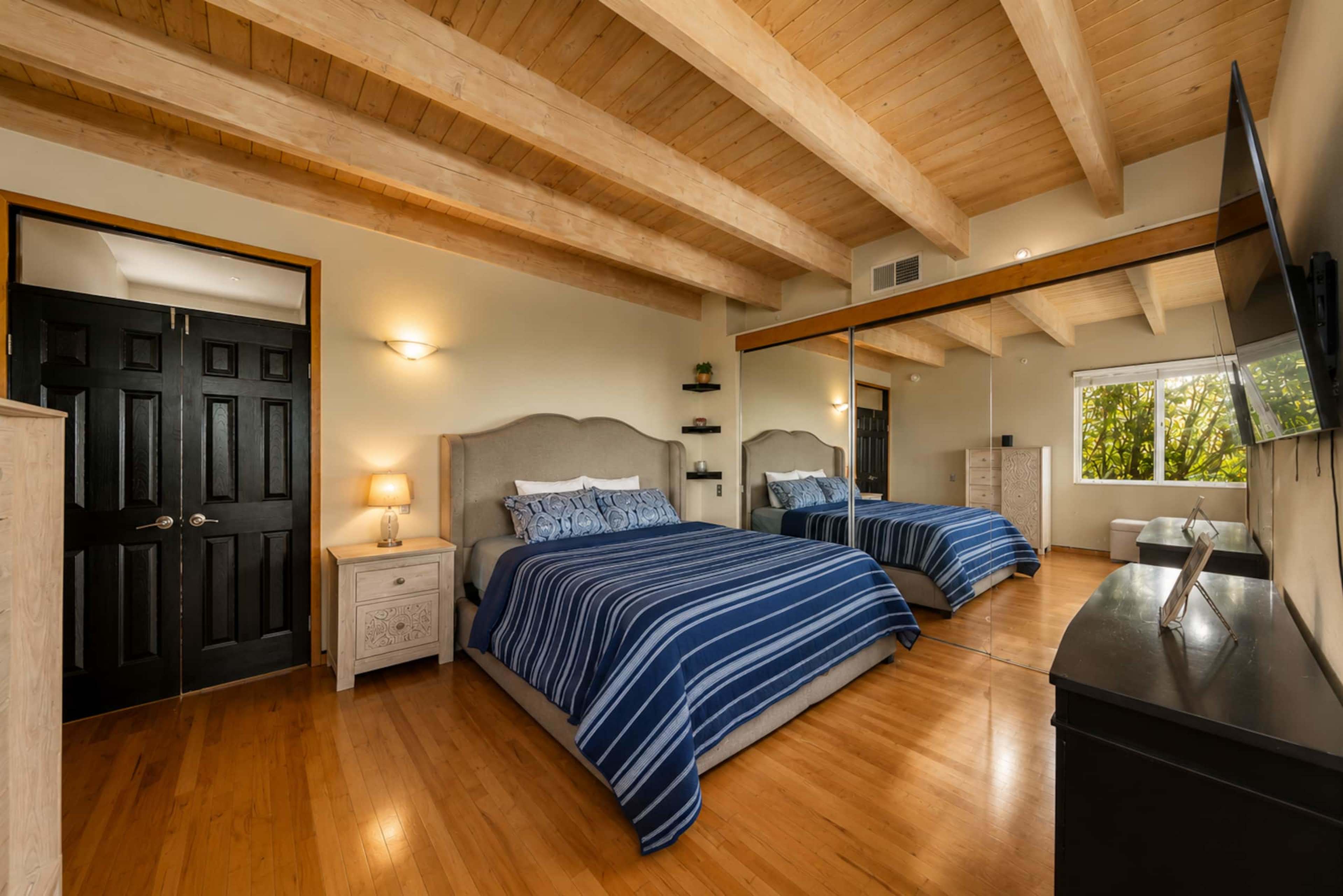 The image shows a spacious bedroom featuring two beds with blue striped bedding, wooden beams on the ceiling, and large windows allowing natural light.