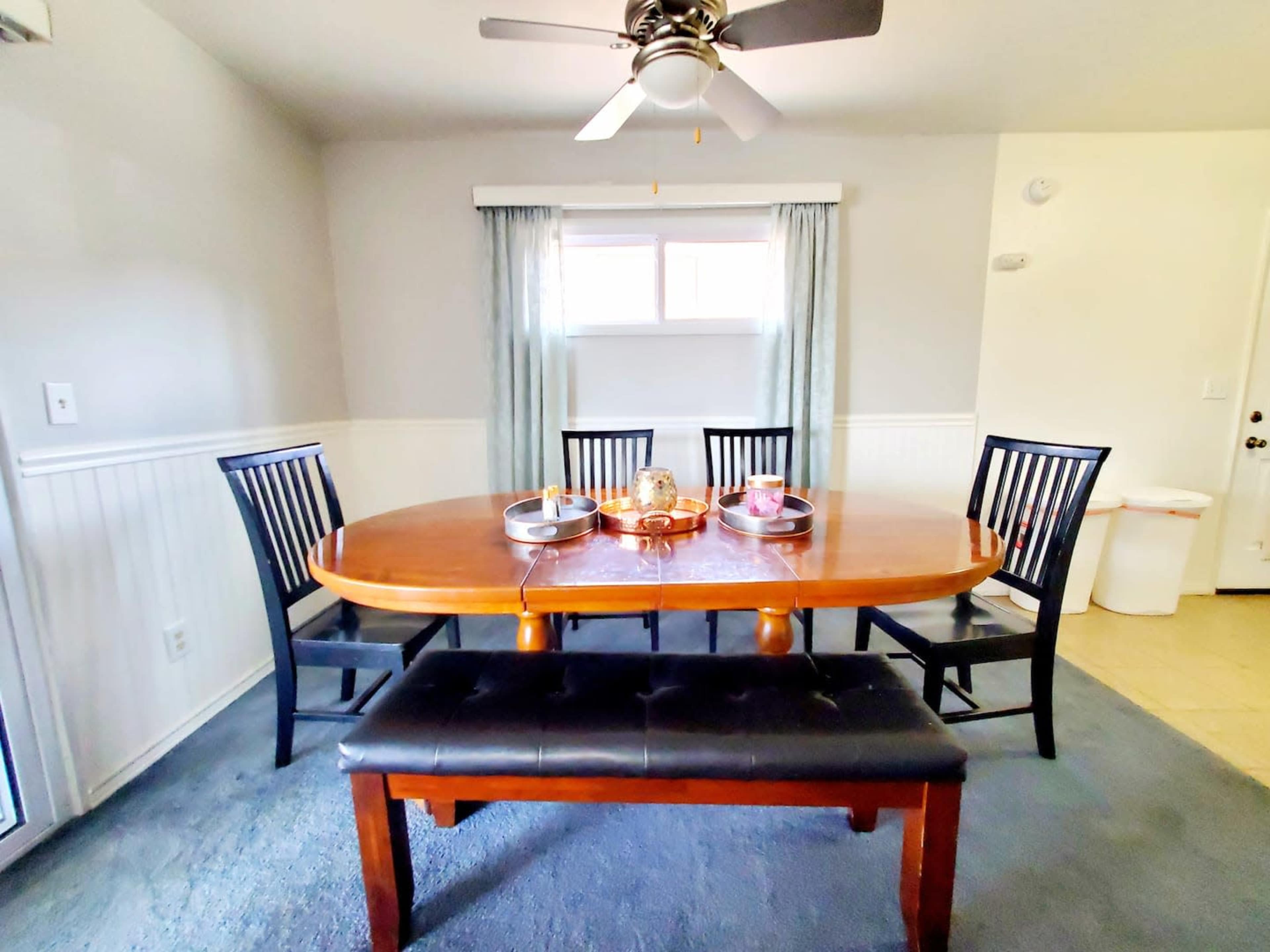 A dining room features a round wooden table set with plates and a centerpiece, surrounded by black chairs and a bench against a light-colored wall.