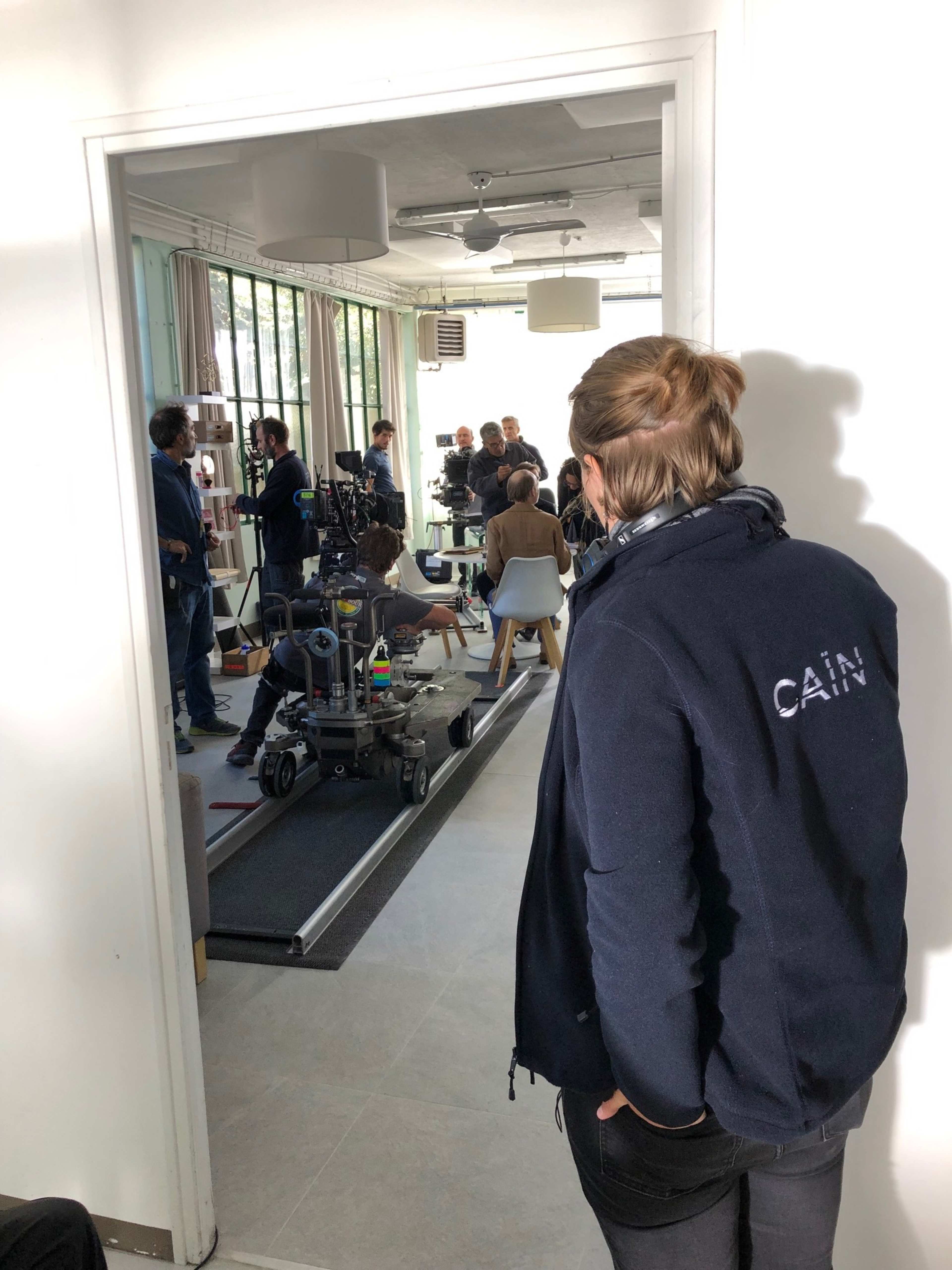 A film crew is set up inside a well-lit room, with cameras and equipment focused on a seated subject, while a person observes from the doorway.