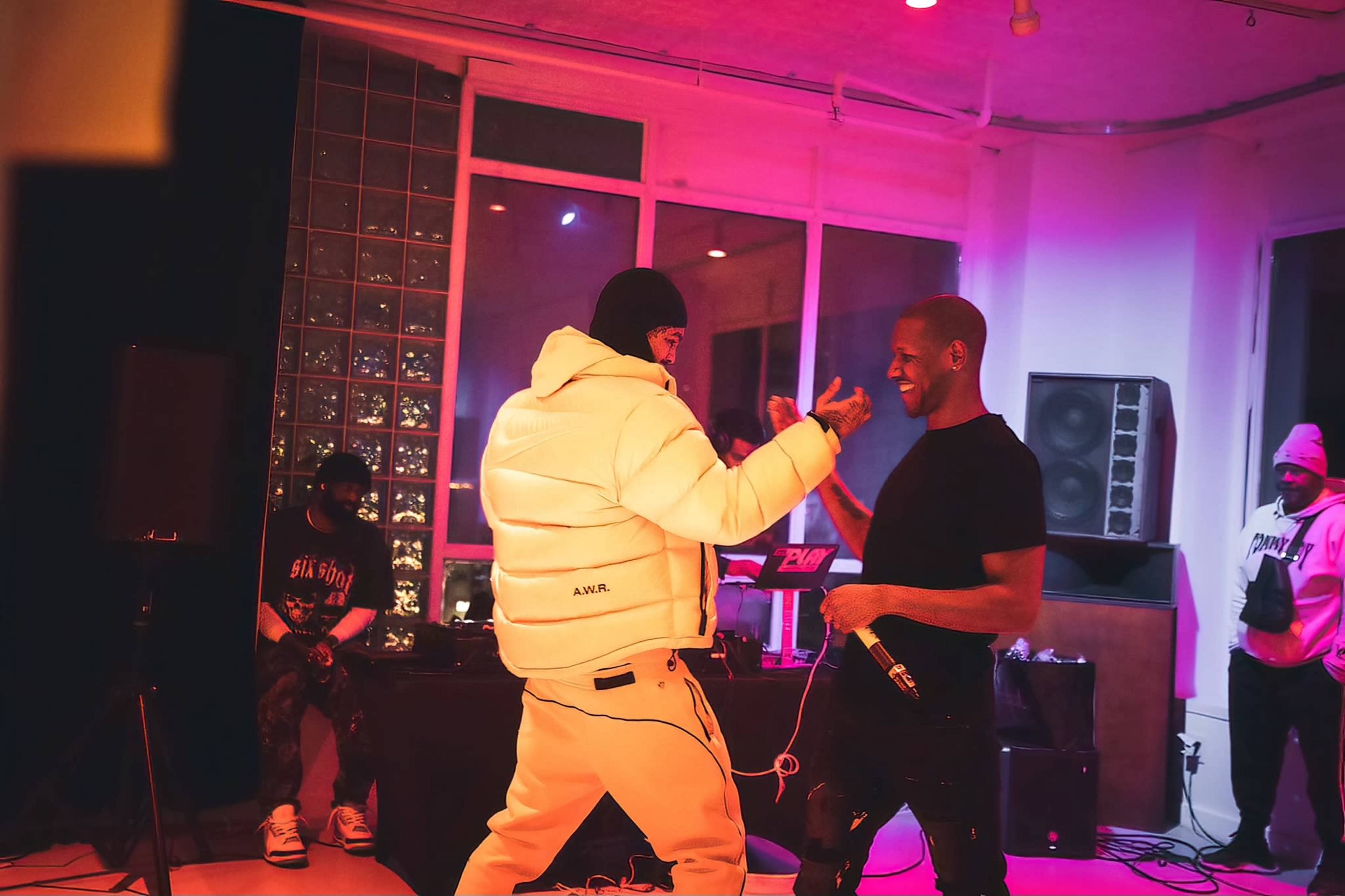 Two performers interact during a live music event in a dimly lit venue, with a DJ and audience members visible in the background.