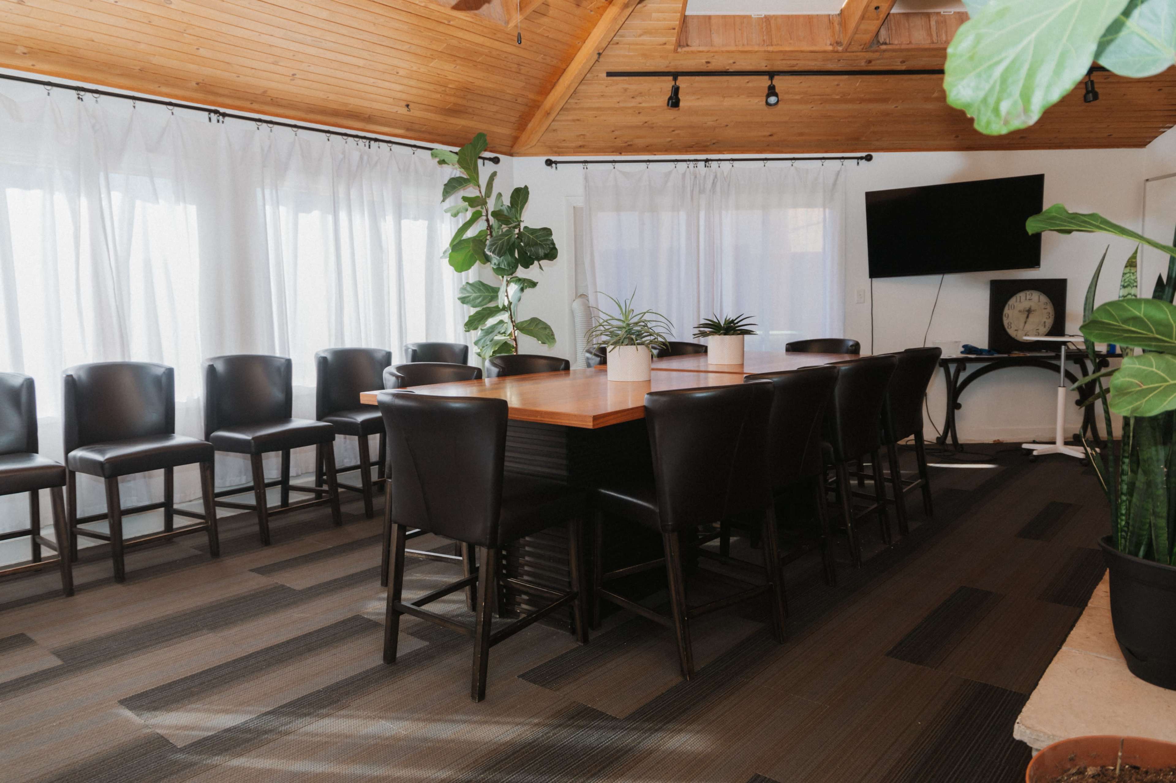 A large wooden conference table surrounded by black chairs is set in a room with high ceilings, large windows, and several indoor plants.