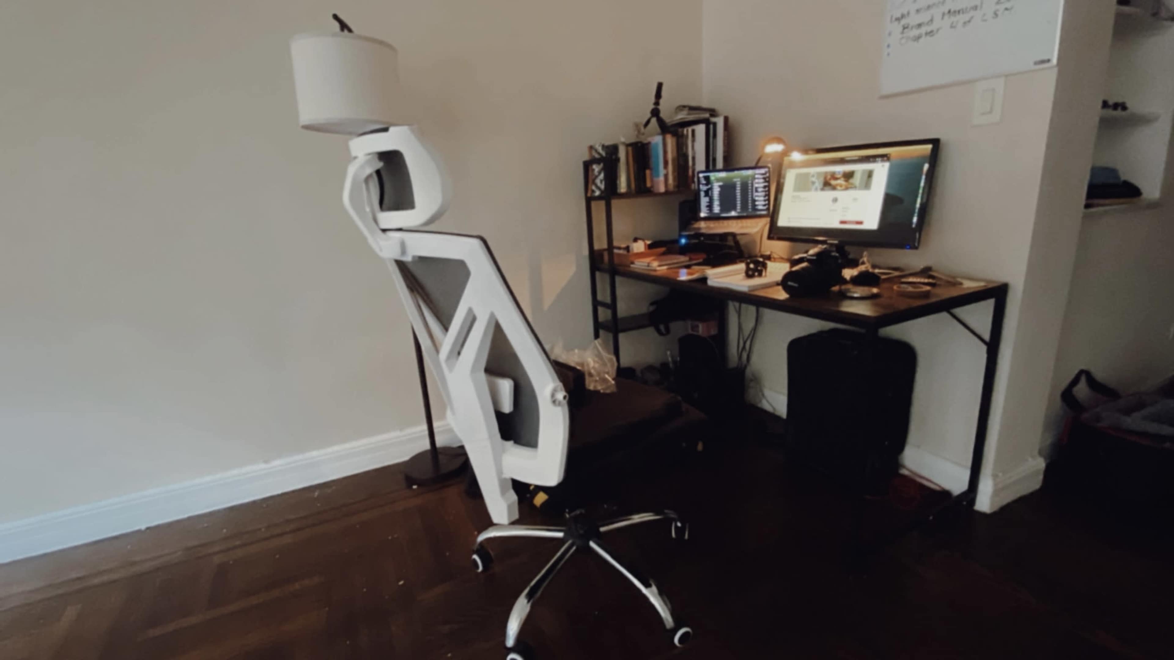 An office setup features a modern chair facing a desk with a computer, lamp, and bookcase against a light-colored wall.