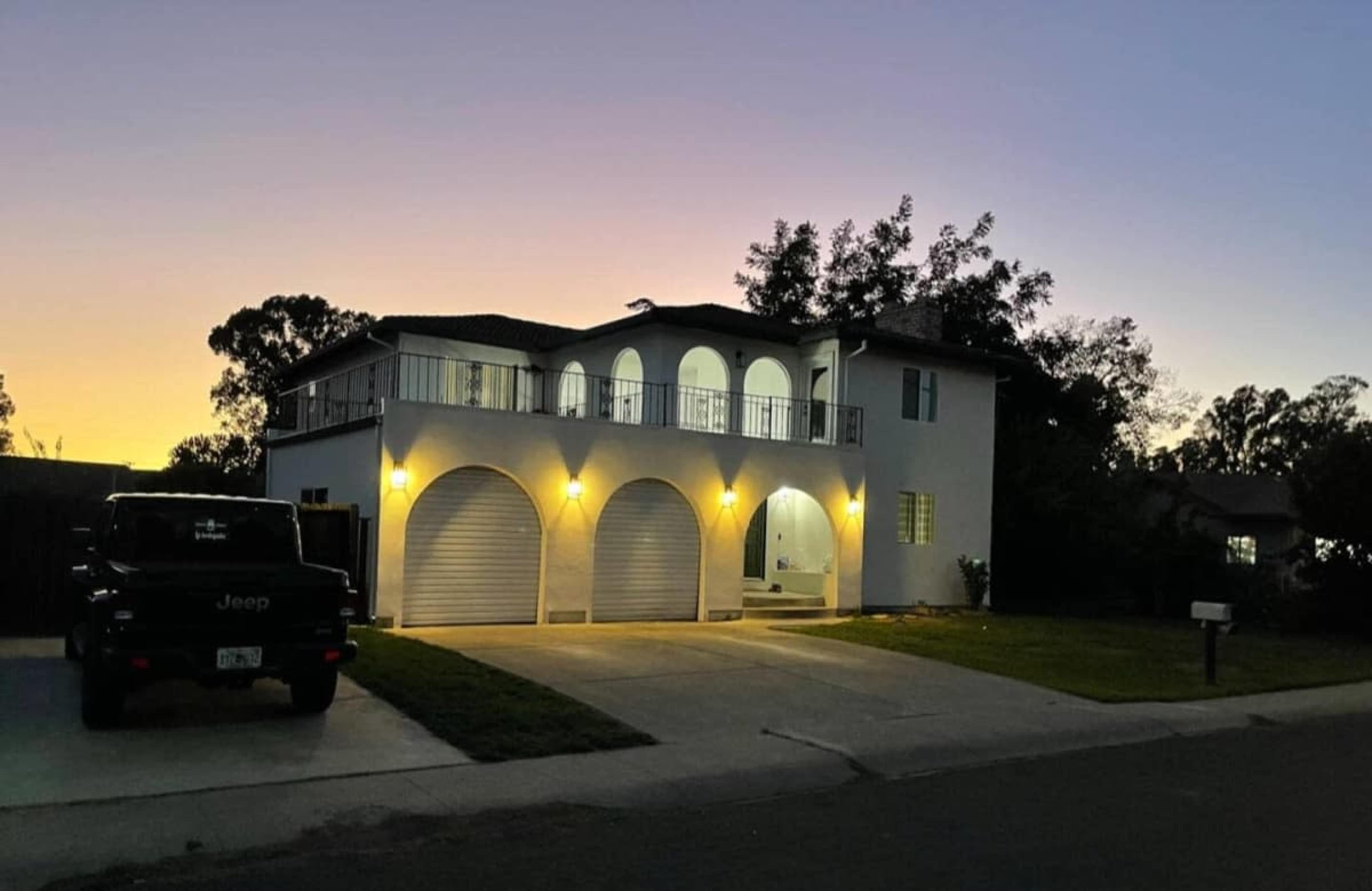 A two-story house features a balcony with arches, three garage doors, and exterior lighting during twilight.