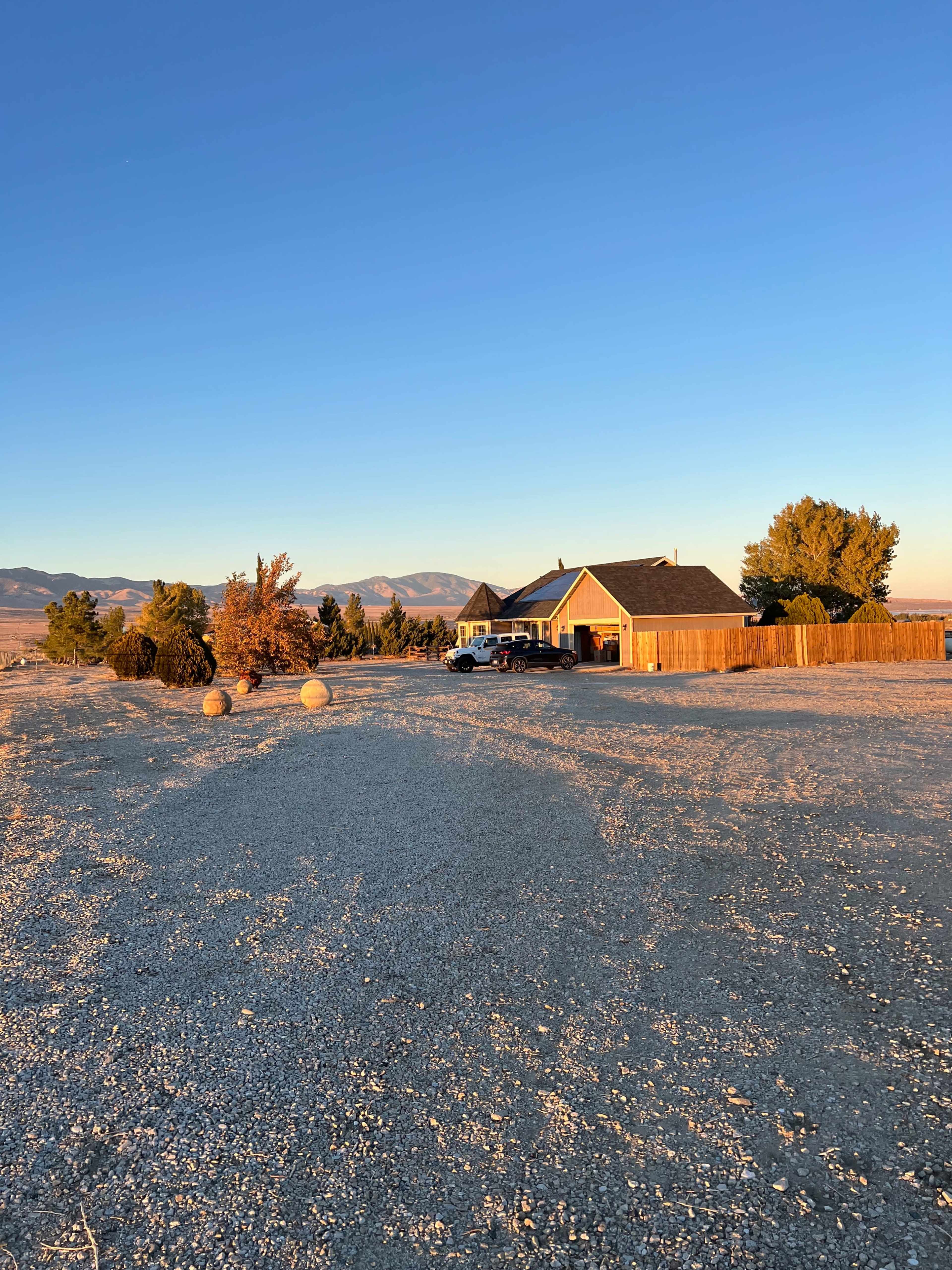 A single-story house is situated on a gravel lot surrounded by trees and distant mountains under a clear blue sky.