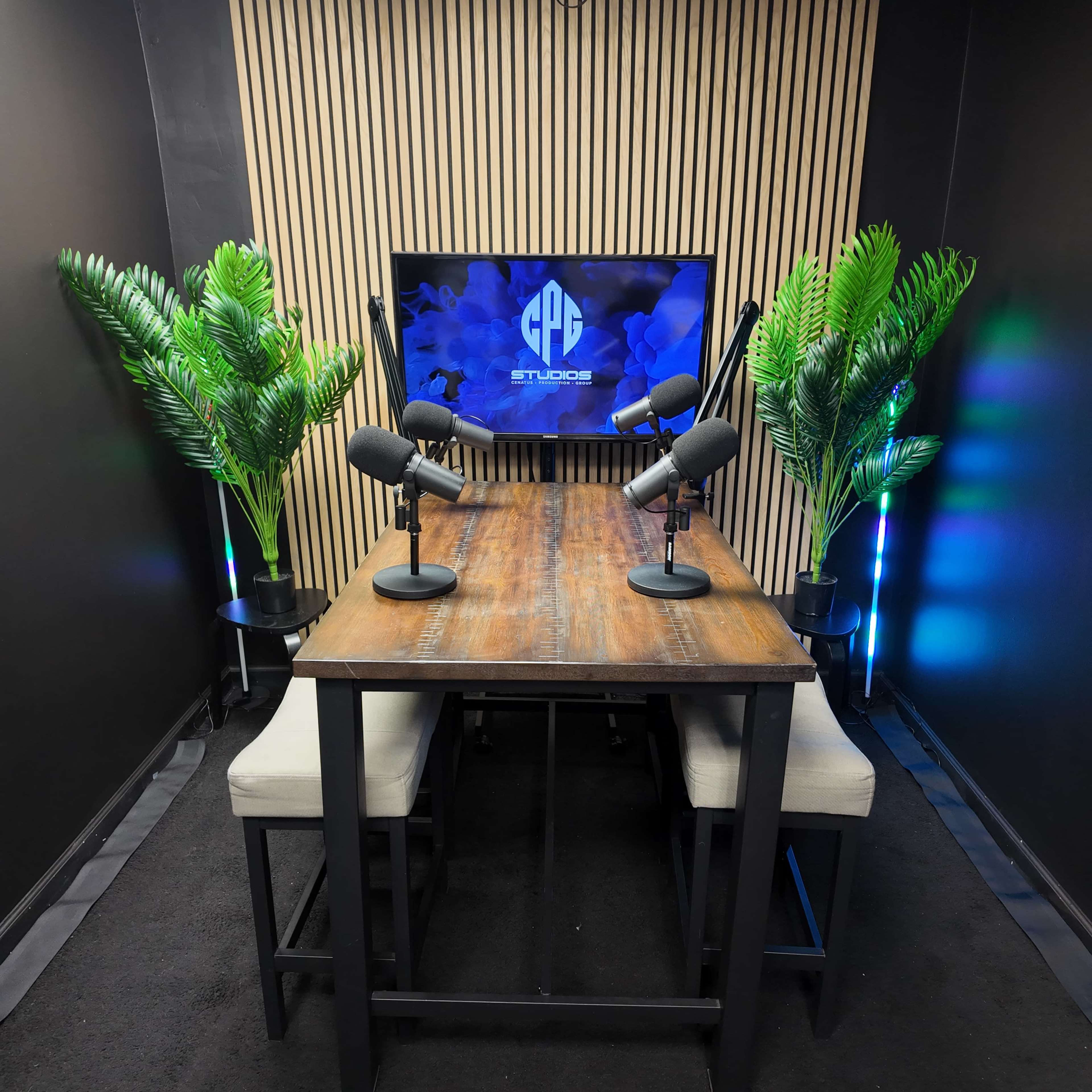 The image shows a podcast studio featuring a wooden table with two microphones, surrounded by tall plants and a screen displaying a logo on a dark wall.