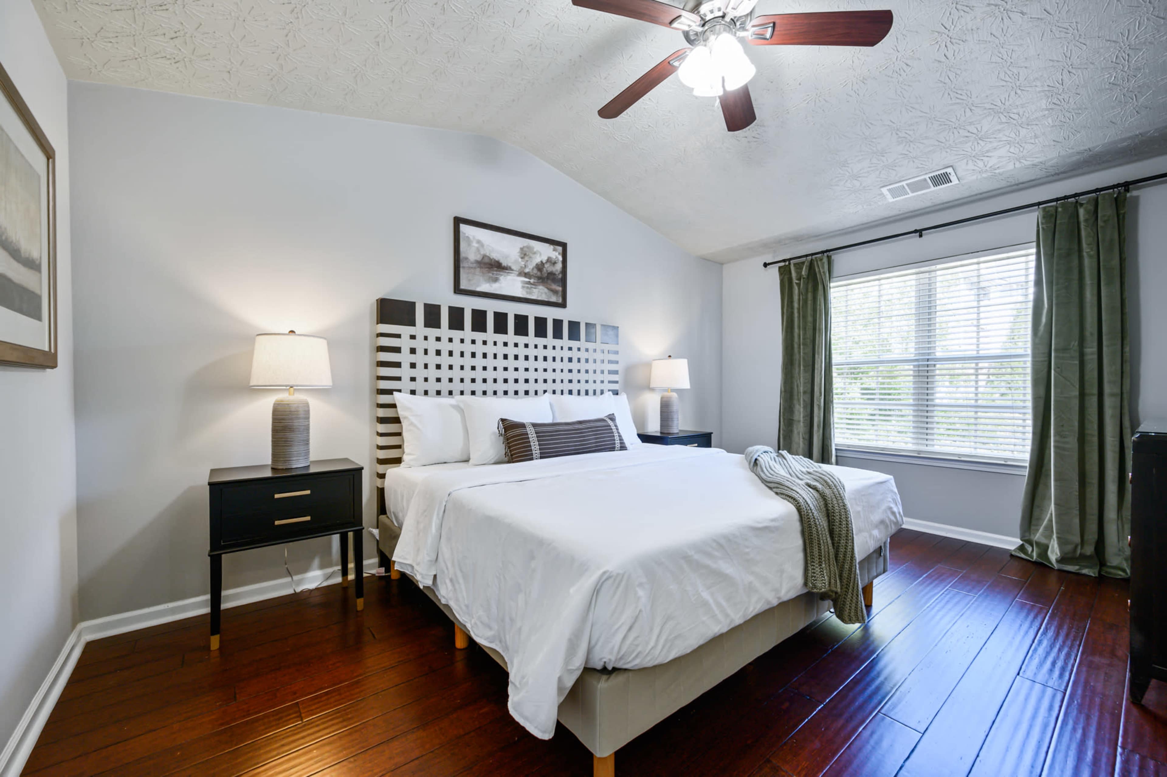 A well-lit bedroom features a king-sized bed with a patterned headboard, two bedside tables with lamps, and large windows dressed with green curtains.