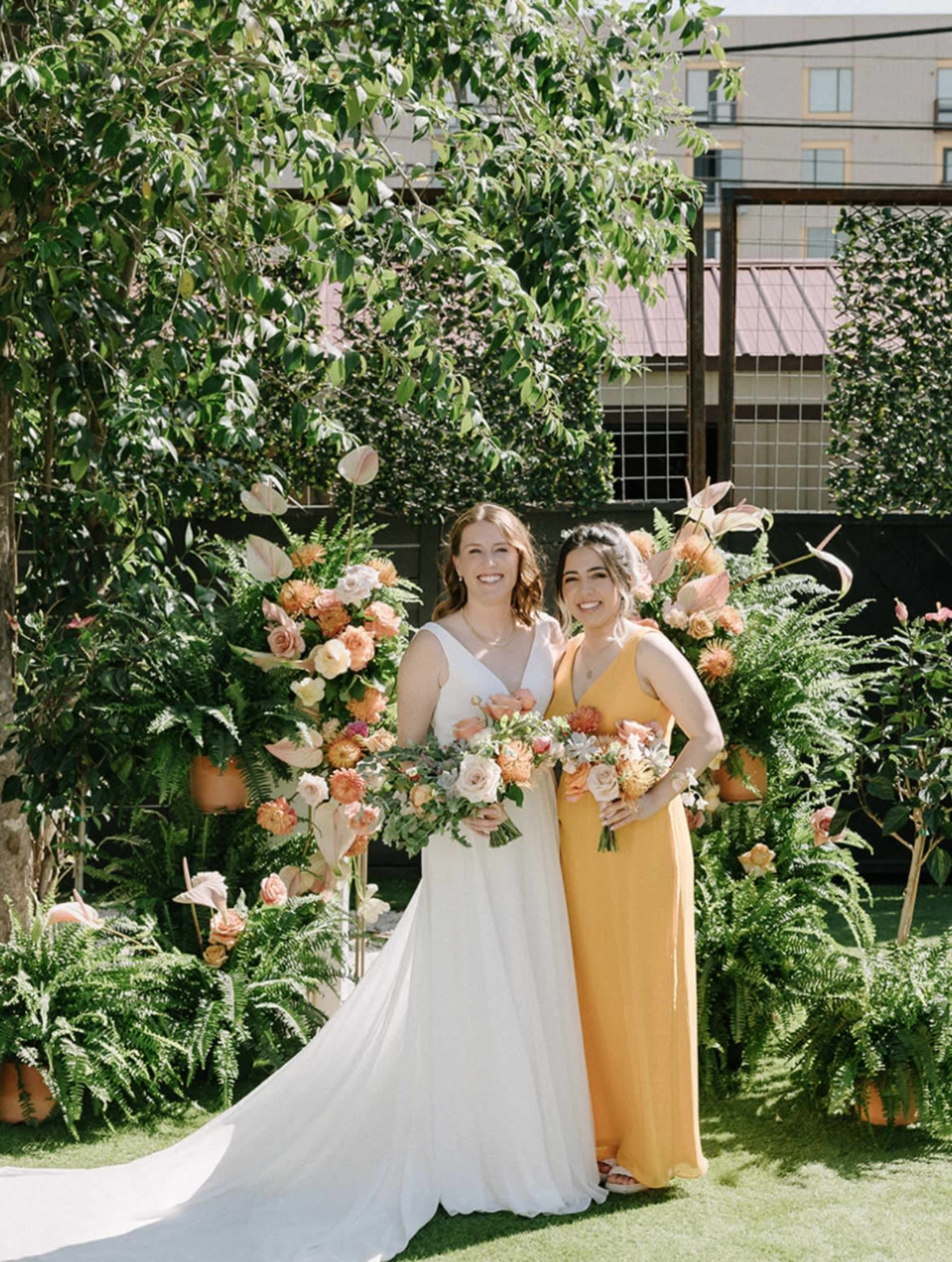 Two women stand smiling in front of a floral backdrop, one dressed in a white wedding gown and the other in a yellow dress, each holding bouquets of flowers.
