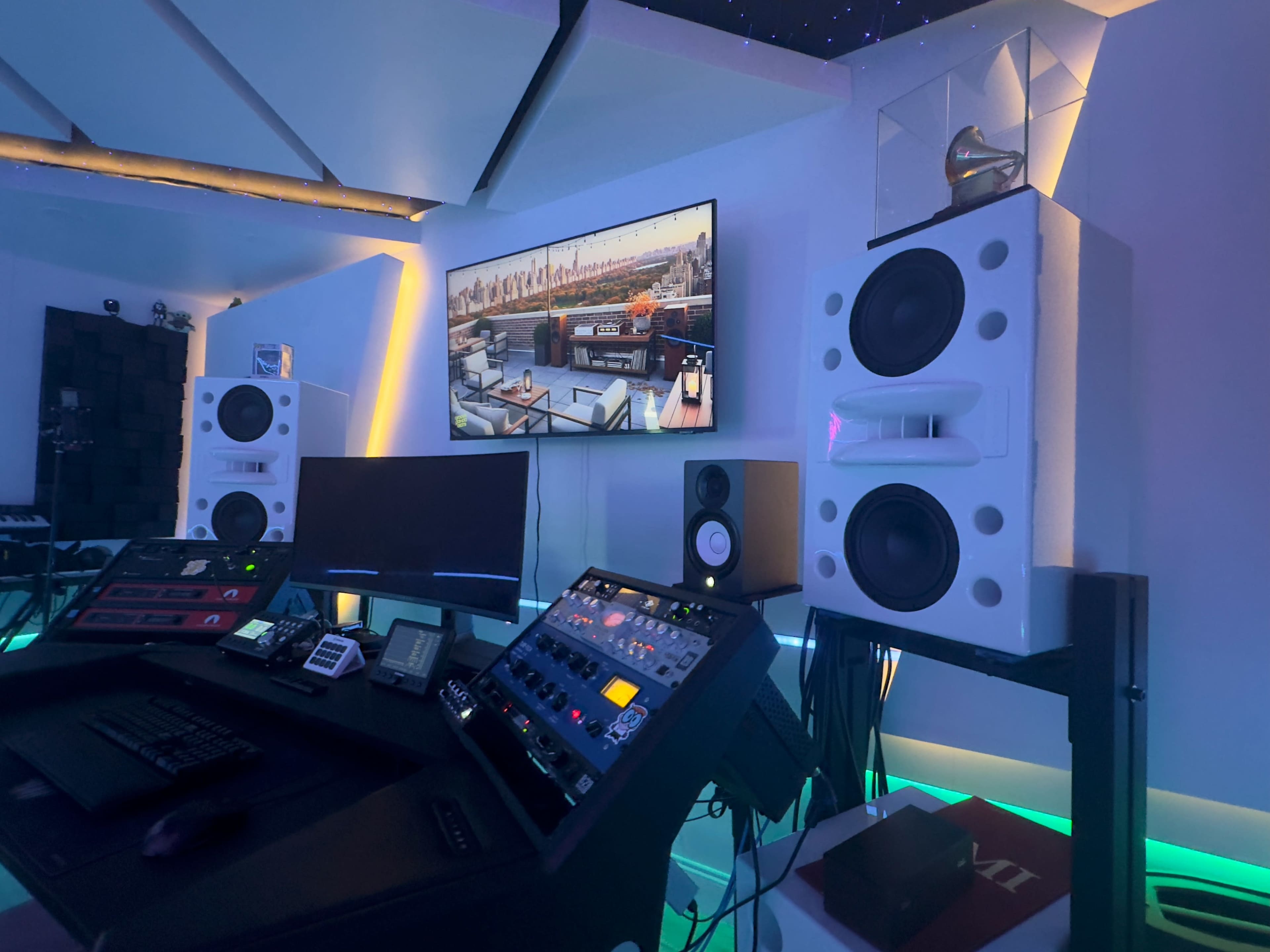 The image shows a modern music production studio with large speakers, a control desk filled with equipment, and a screen displaying a cityscape.