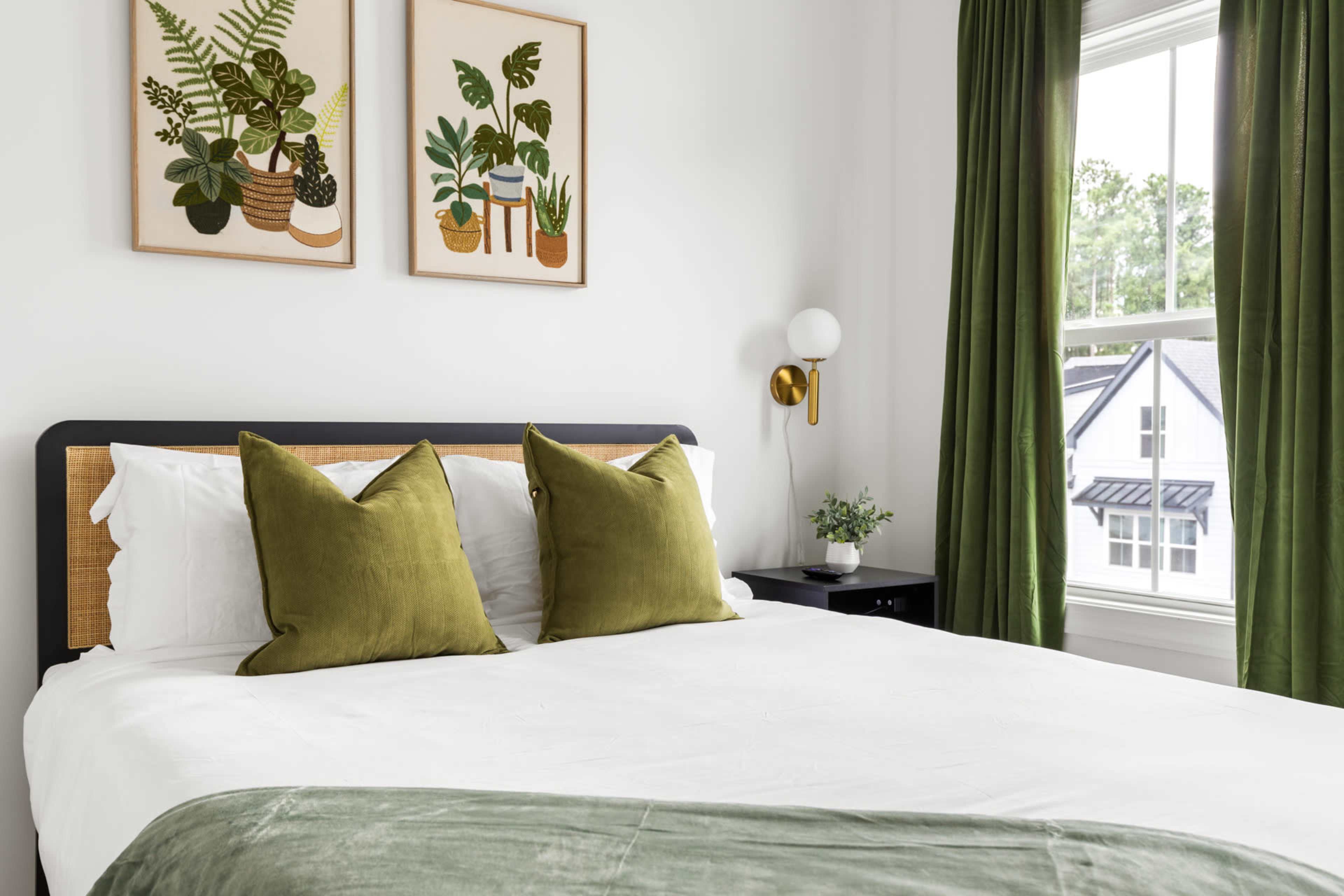 A neatly made bed with green decorative pillows is positioned in a bright room featuring plant-themed artwork and green curtains.