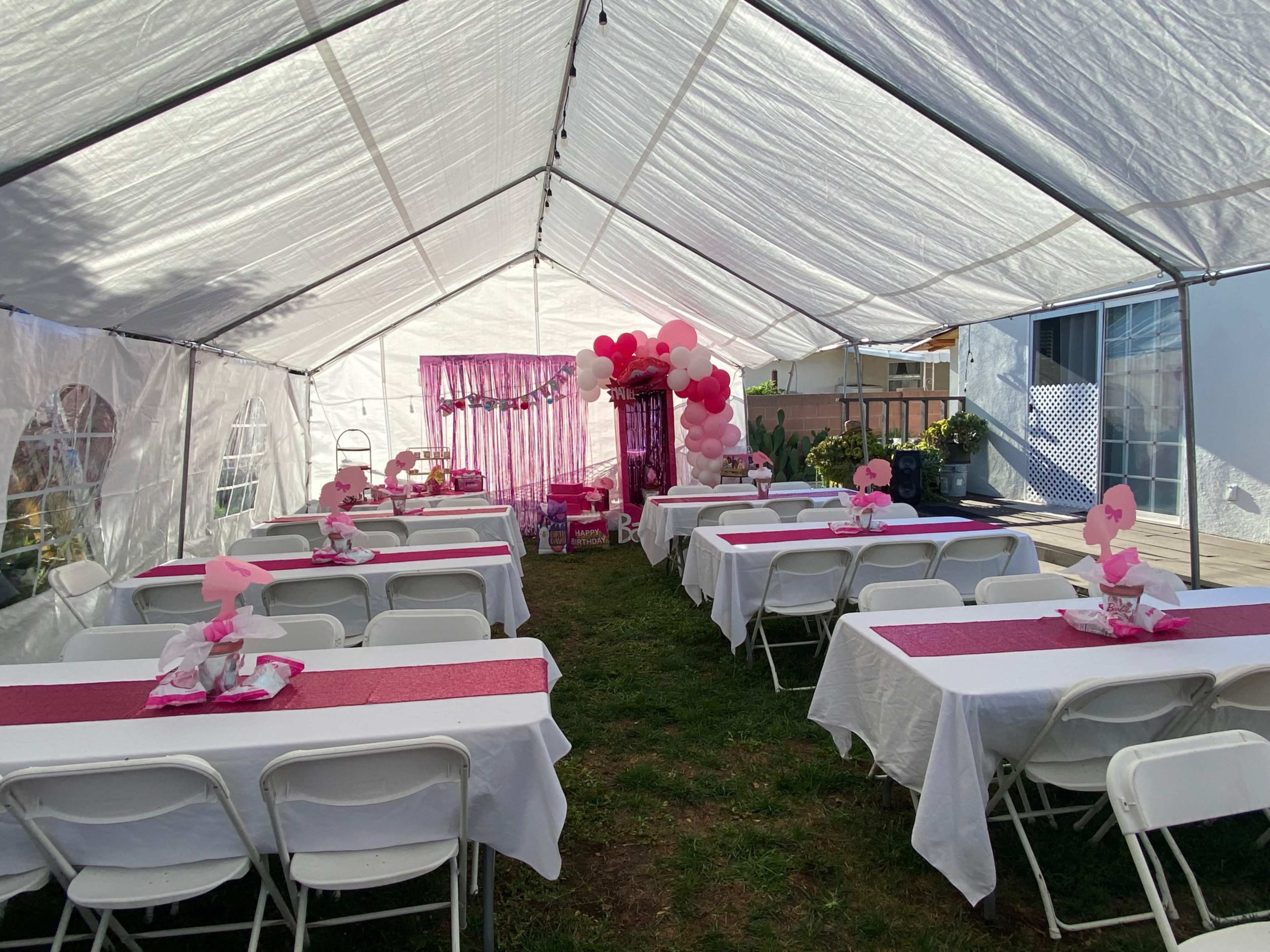 The image shows a decorated outdoor tent with rows of white tables covered in pink tablecloths, featuring floral centerpieces and balloon decorations.