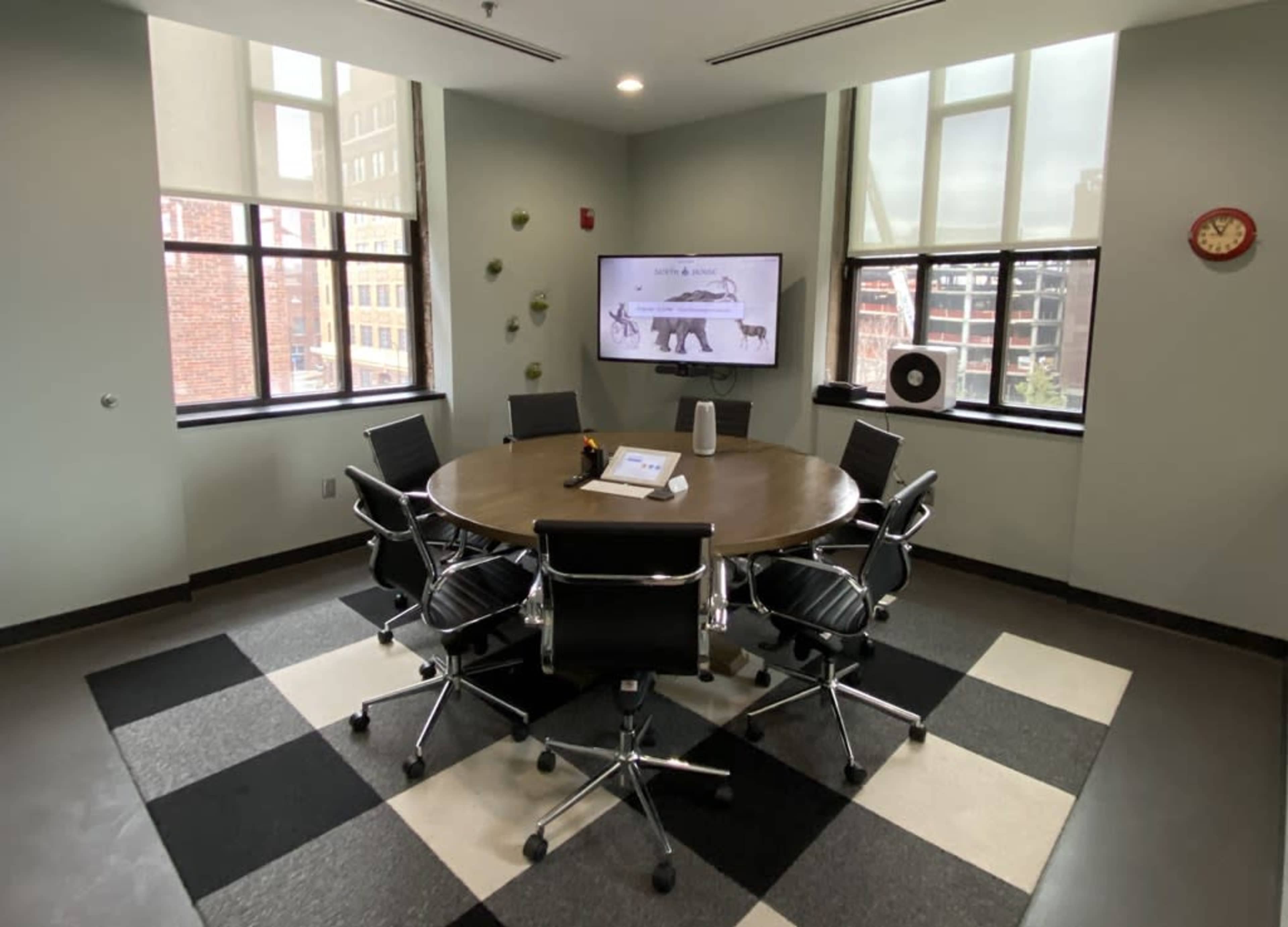 The image shows a modern conference room with a round wooden table surrounded by black leather chairs, a television displaying an image of bears, and large windows allowing natural light.