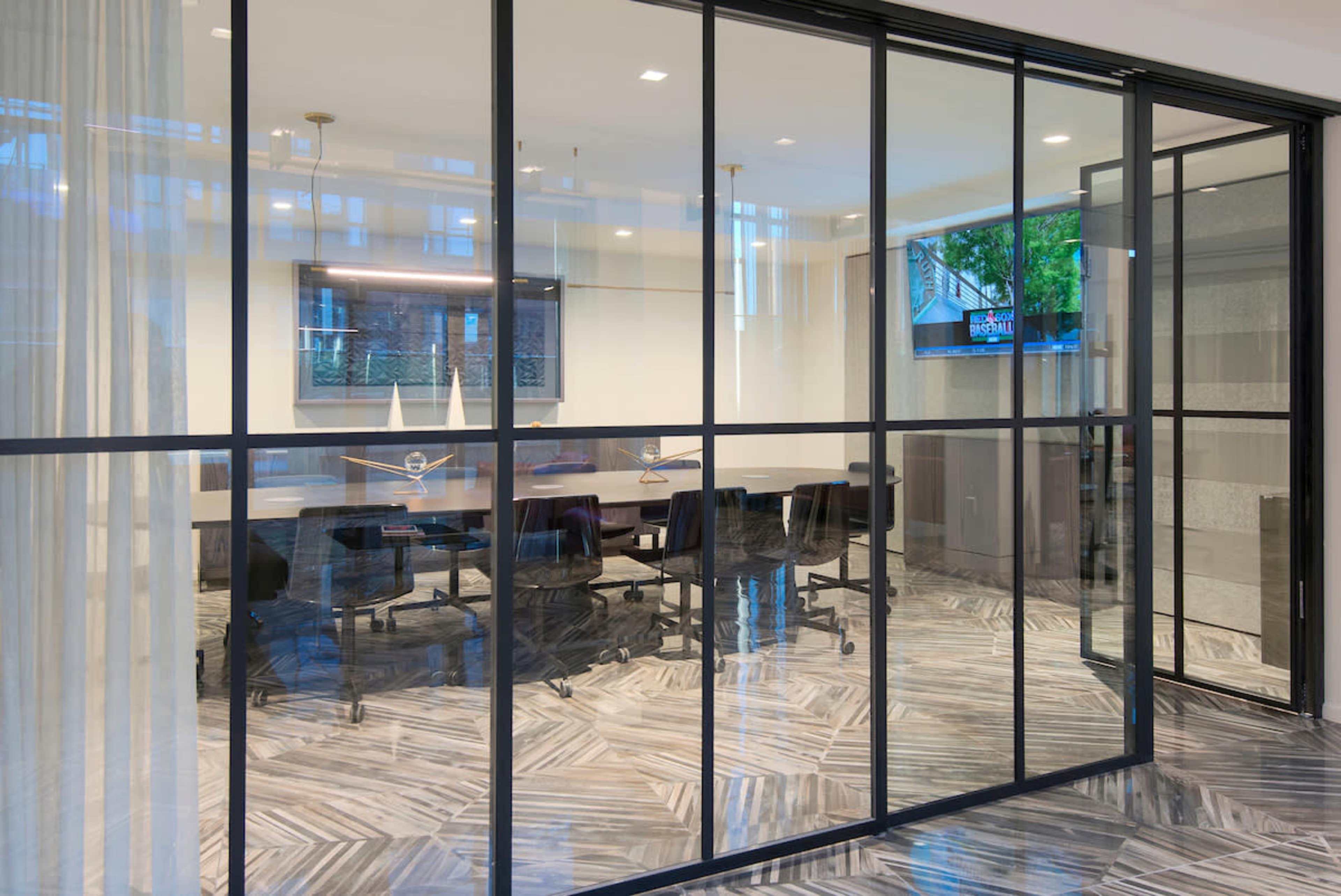 A modern glass-walled conference room with a large table and office chairs, featuring a television mounted on the wall.