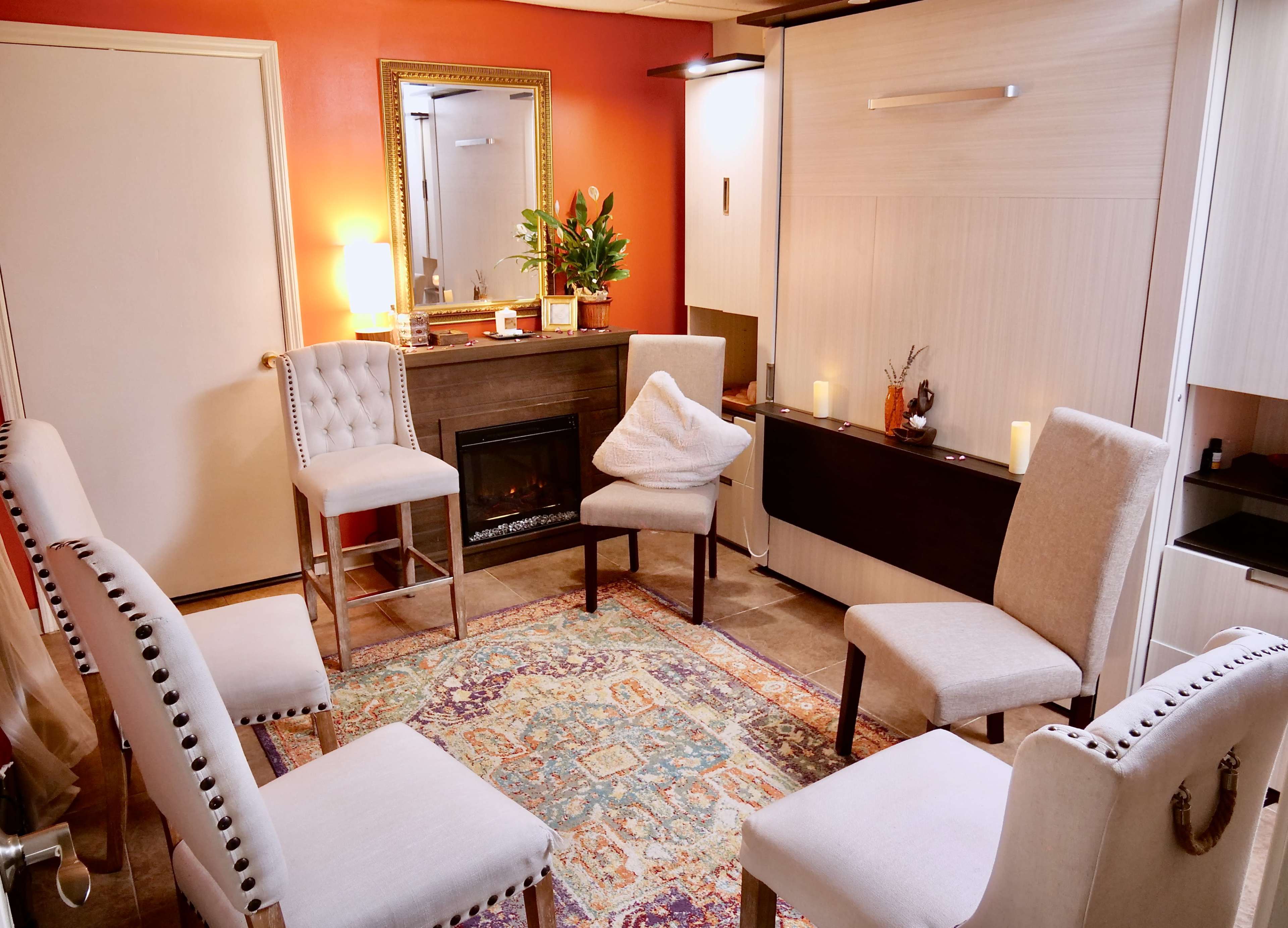 A cozy room features a circular arrangement of upholstered chairs around a decorative rug, with a small electric fireplace and a potted plant against an orange accent wall.