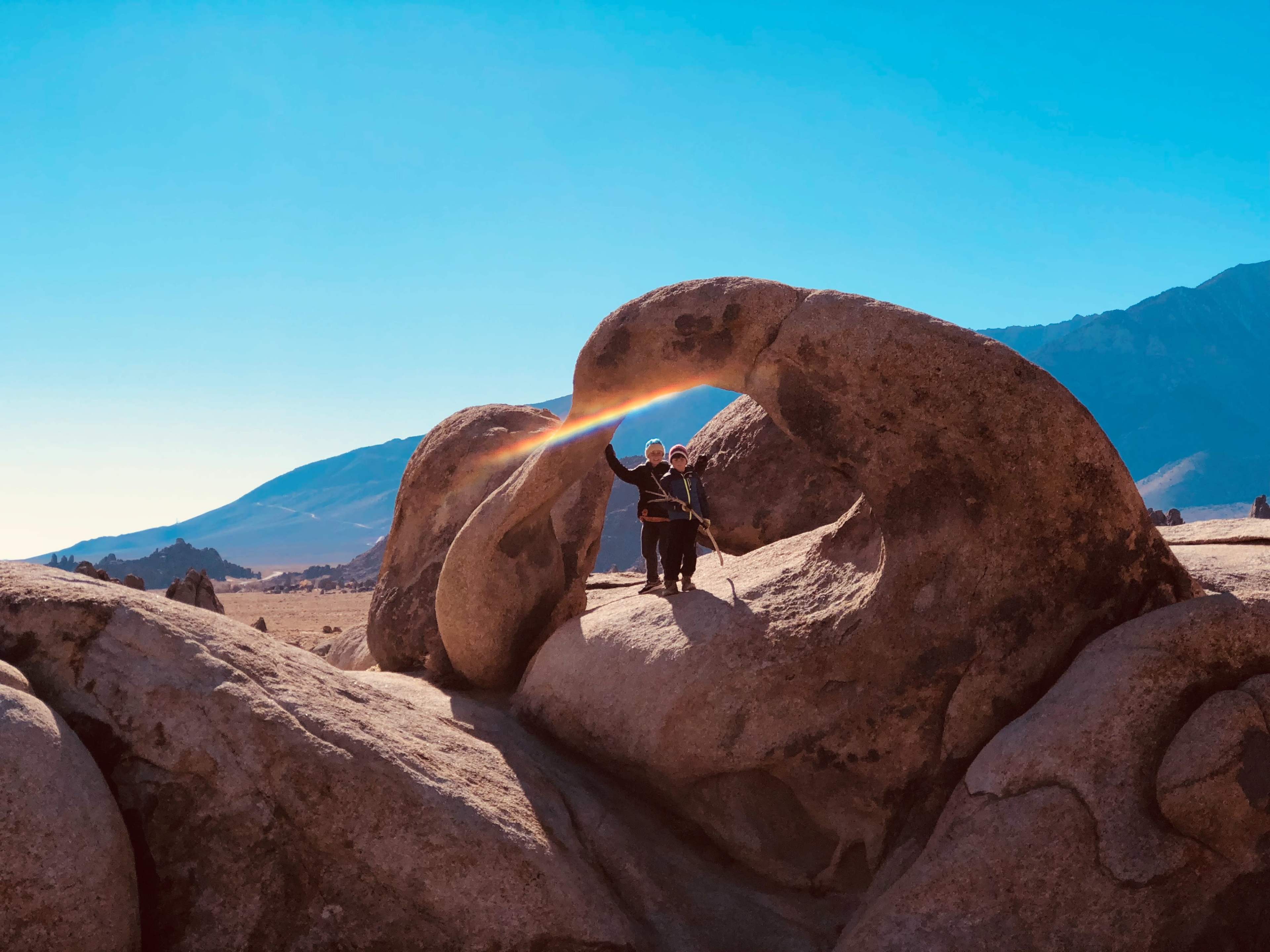 Two people stand under a natural rock arch with a rainbow visible in the opening against a clear blue sky.