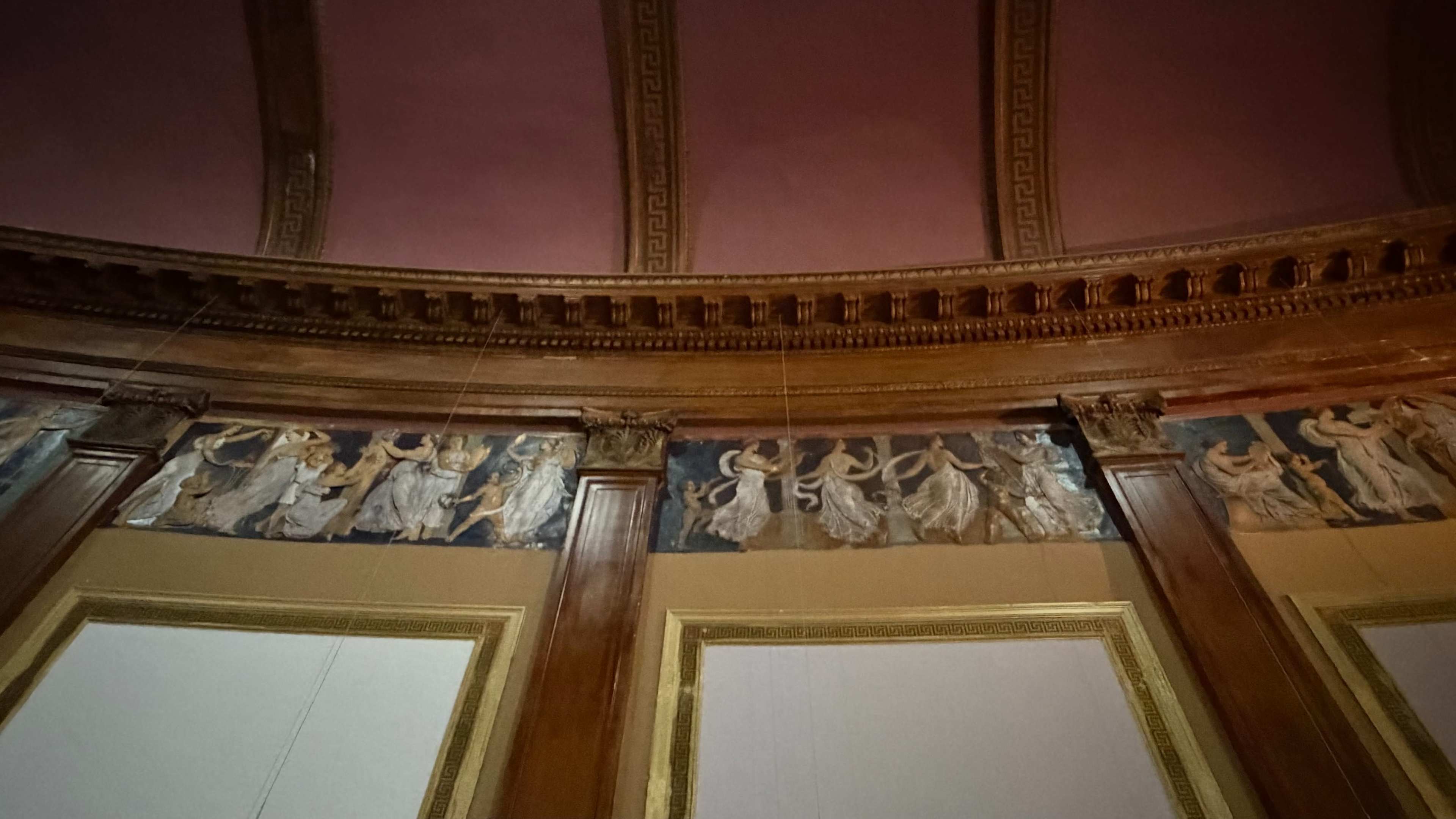 The image shows a circular room featuring a decorative frieze with figures in flowing robes, set against a dark background, beneath a wooden cornice and a pink ceiling.