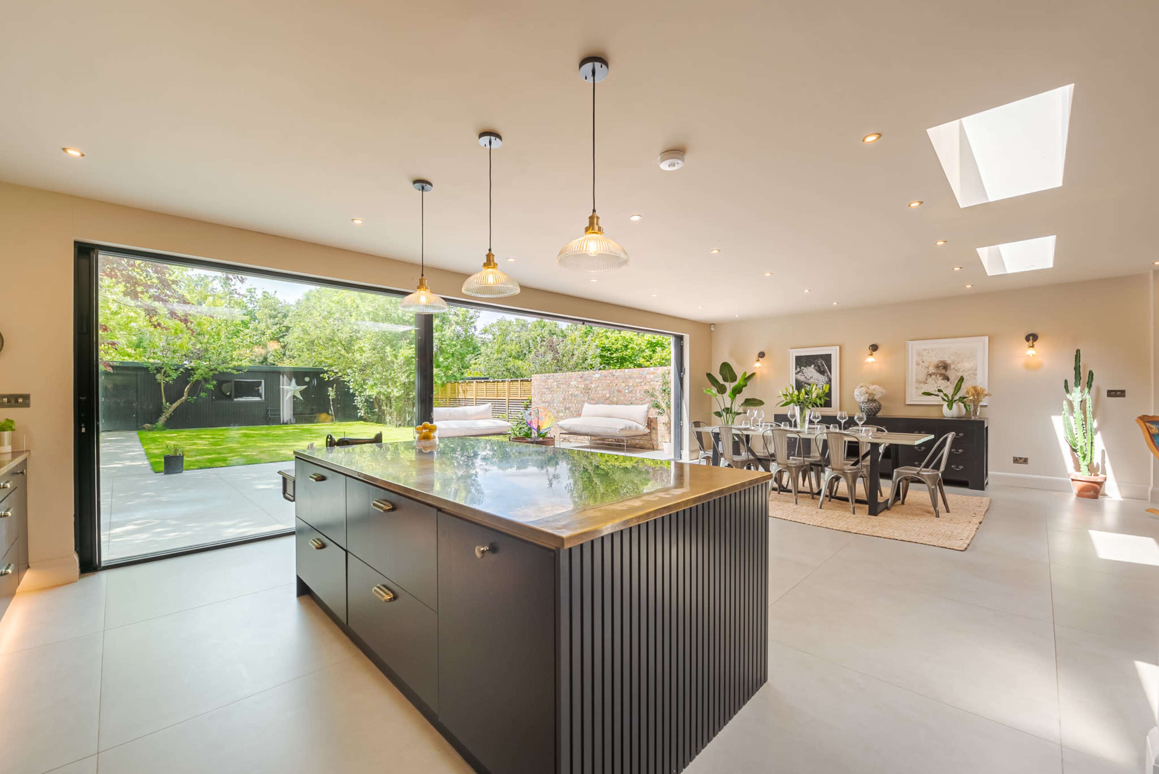 The image shows a modern kitchen and dining area with large glass doors opening to a garden, featuring an island with a stone countertop and a dining table set for six.