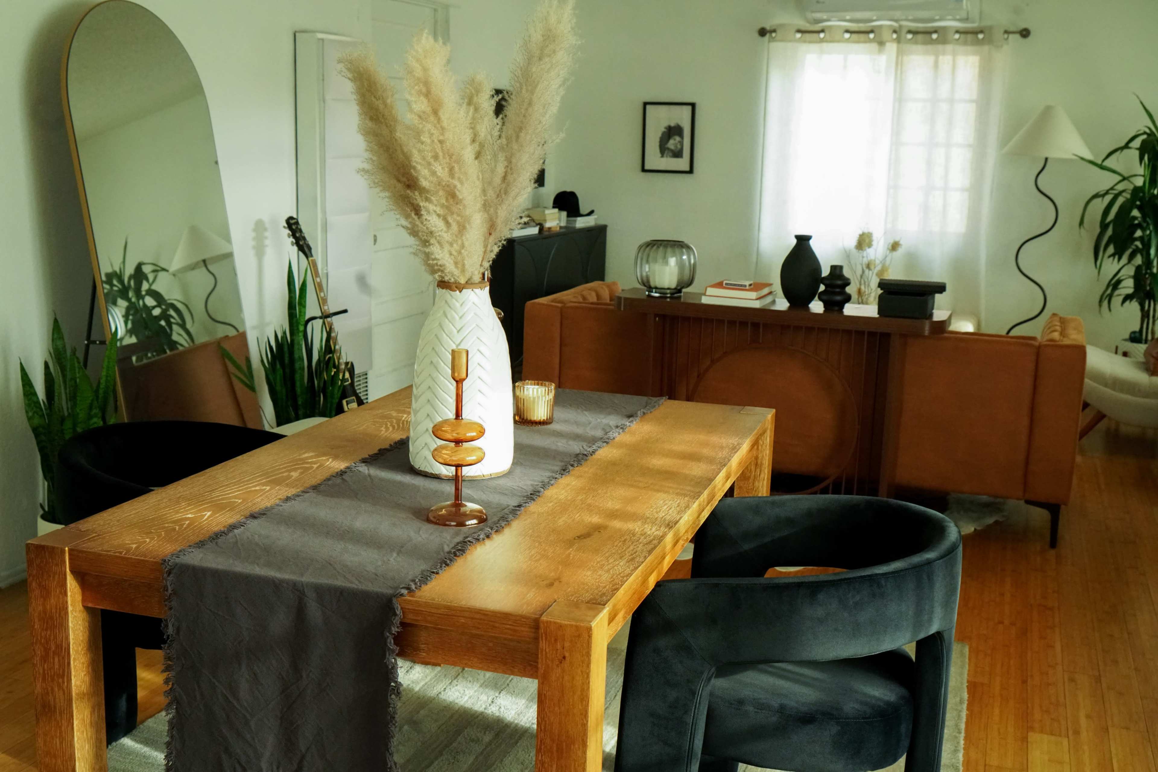 The image shows a modern living space featuring a wooden dining table with a decorative vase and a candle, surrounded by dark chairs and illuminated by natural light.